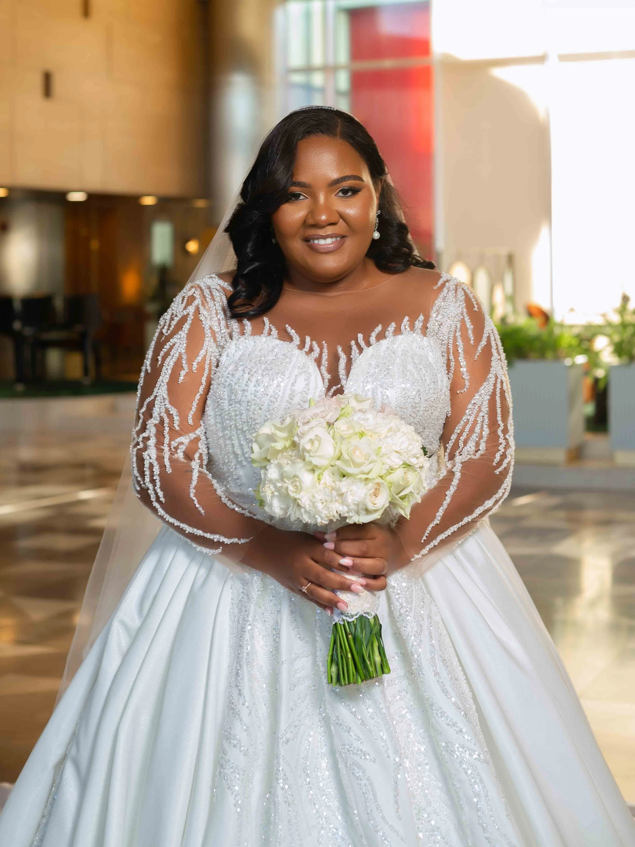 Mulher sorridente vestida de noiva segurando um buquê de flores brancas, com cabelo escuro e vestido branco com detalhes de brilhos, em ambiente interno iluminado.