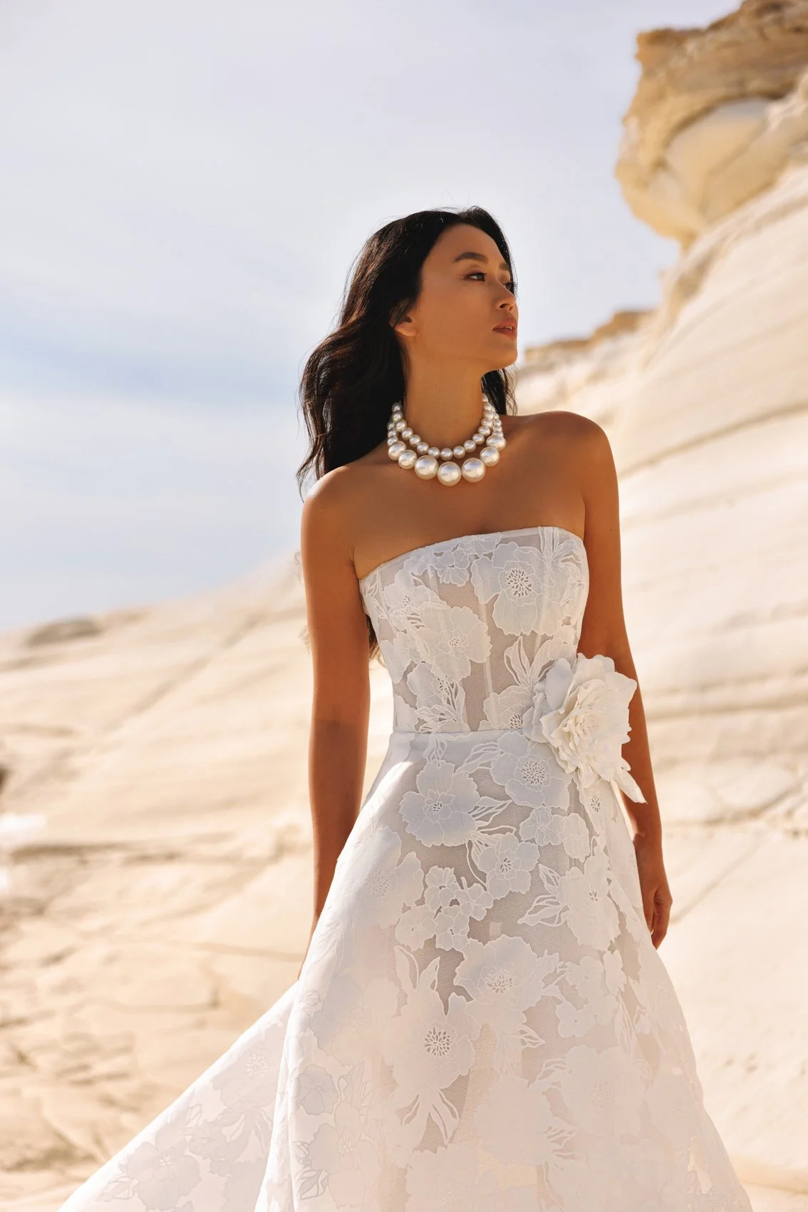 Uma mulher em um vestido de renda branco sem alças com padrões florais e um grande detalhe de flor na cintura, de pé ao ar livre contra uma paisagem rochosa, deserto-like com um céu claro, vestindo um colar de pérolas multi-strand.