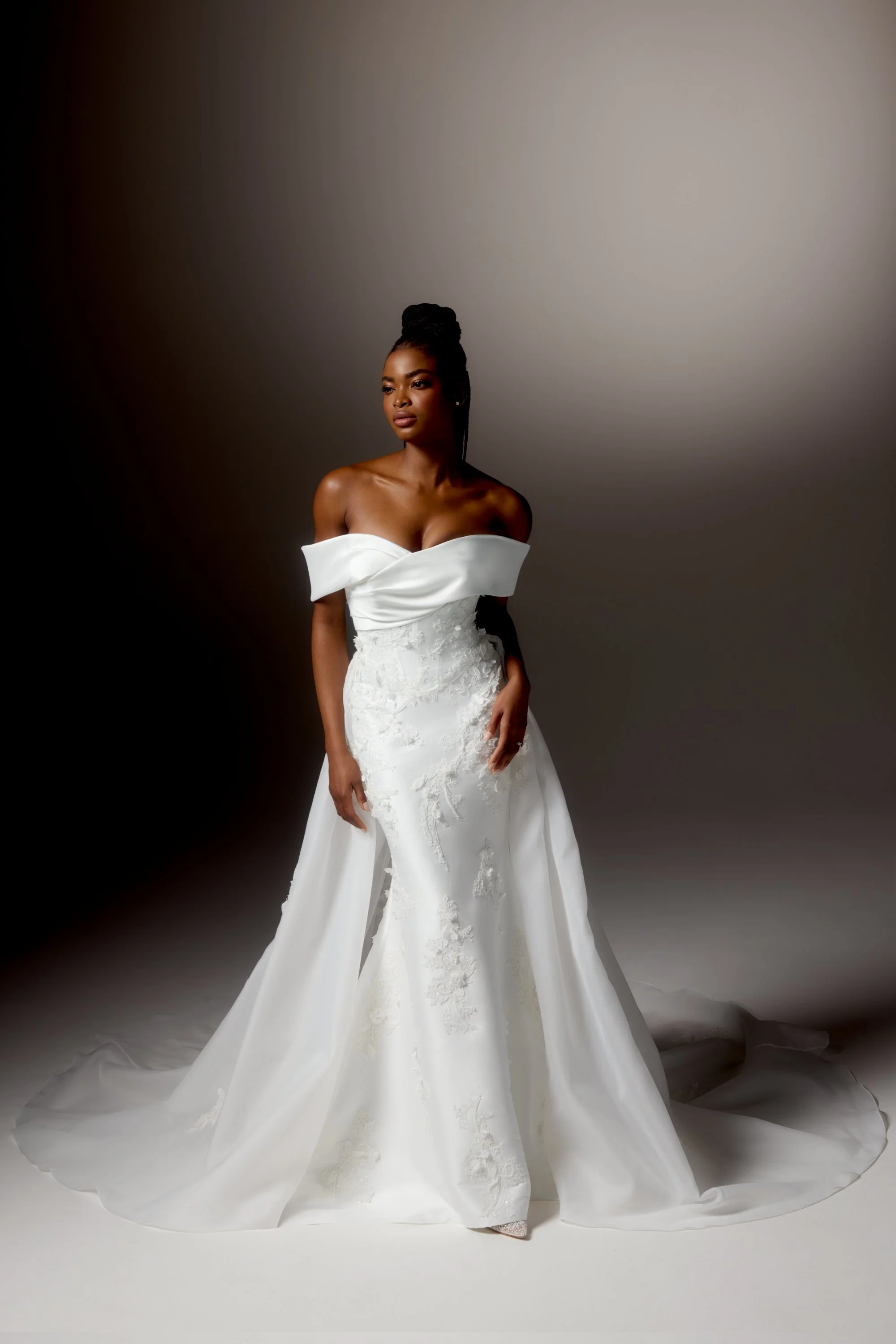 Mulher em vestido de noiva branco com detalhe de rendas, de cabelo trançado, posando contra fundo cinza