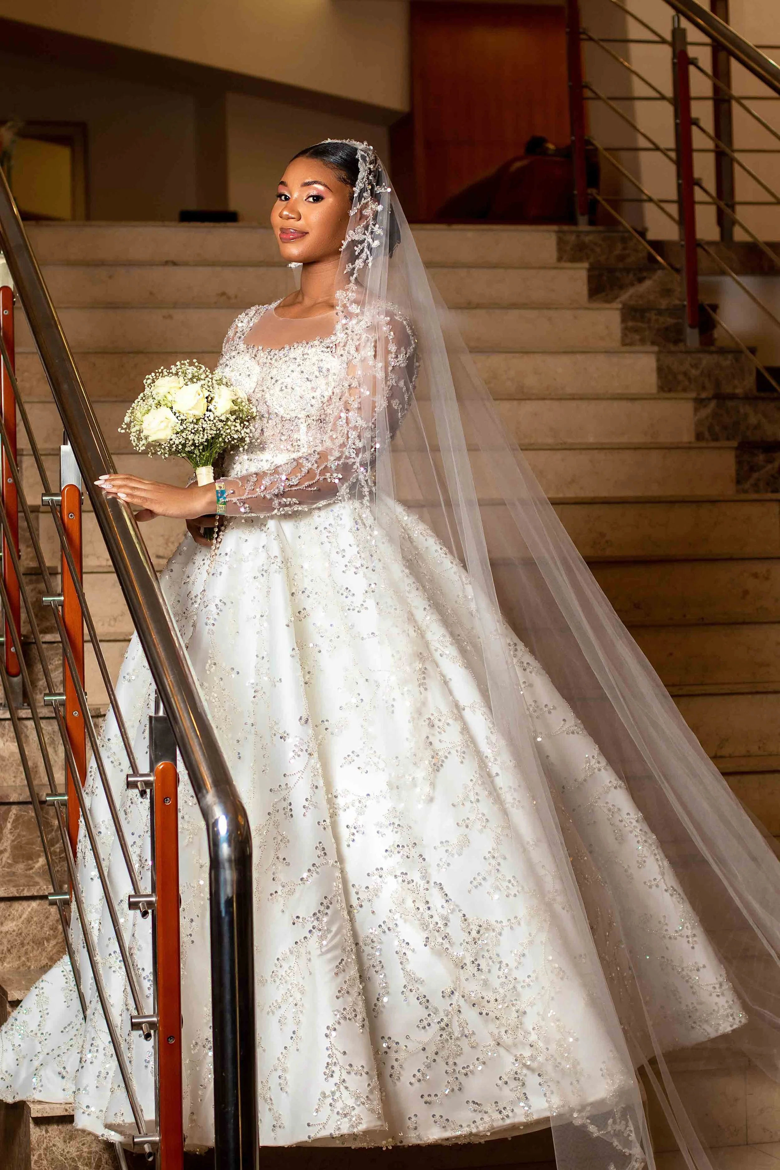 Noivas de vestido branco com véu, segurando um buquê de flores brancas, subindo uma escada de marfim com detalhes brilhantes.