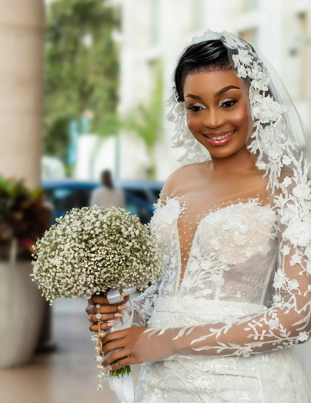 Noiva sorridente usando vestido de noiva com véu de renda, segurando buquê de flores pequenas brancas, em fundo ao ar livre com árvores e edifícios.