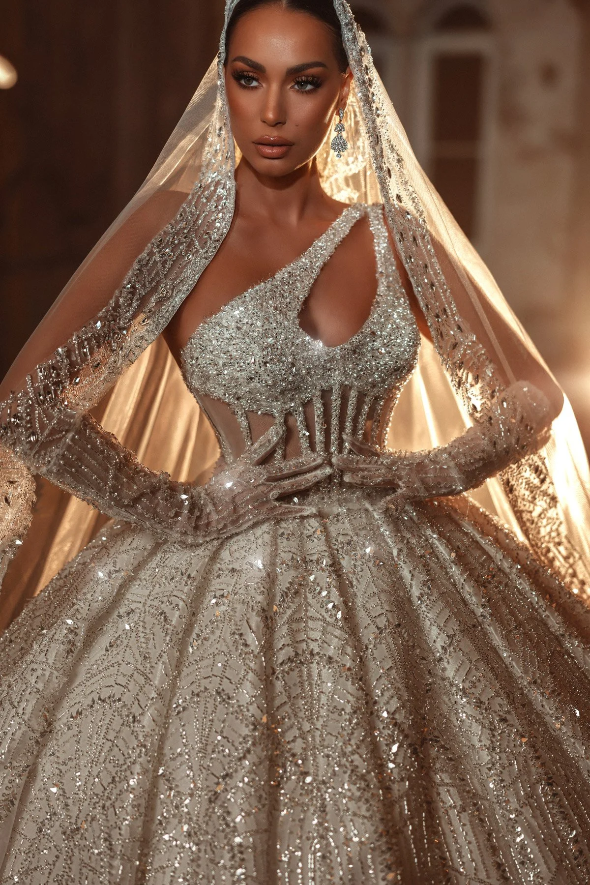 Mulher vestida com vestido de noiva em tom dourado, decorado com detalhes de pedreria e glitter, usando véu e brincos sofisticados.