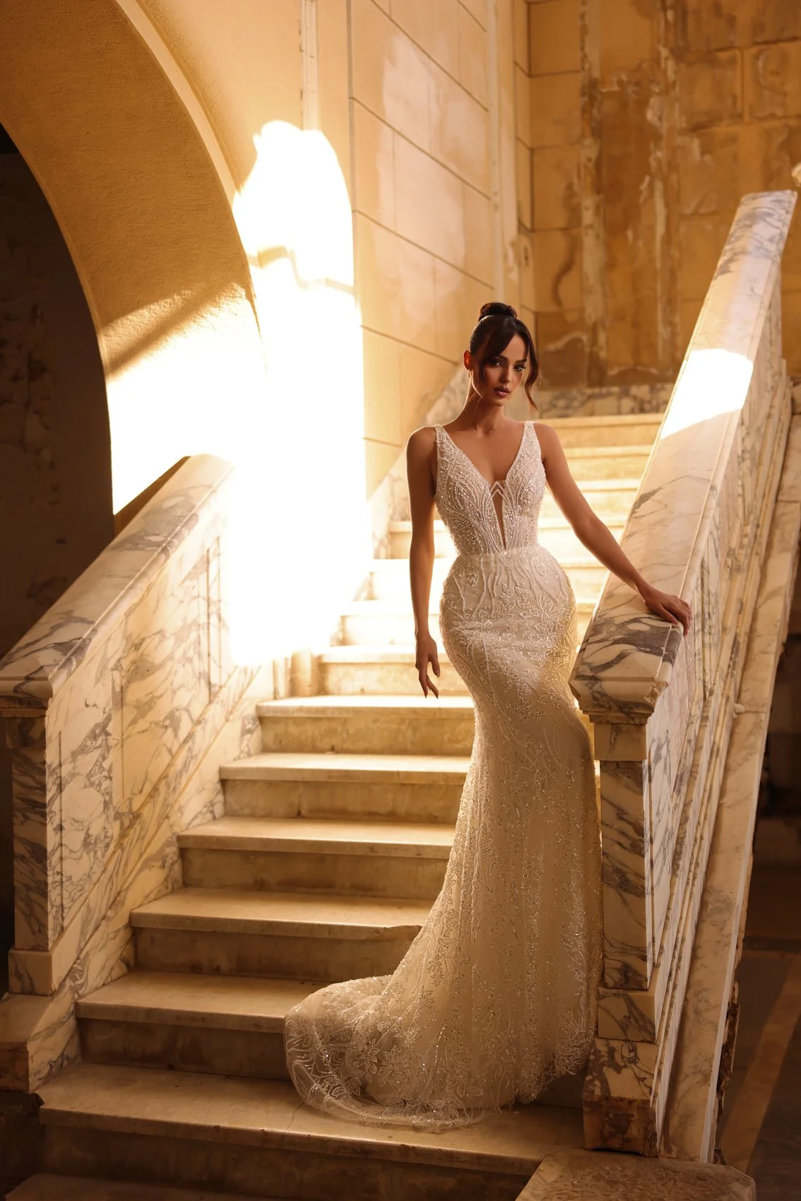 Uma mulher em um elegante vestido de noiva de renda branca de pé em uma escada de mármore iluminada por iluminação quente.