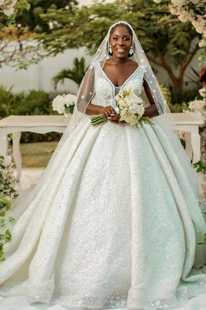 Noiva usando vestido de casamento branco e véu, segurando um buquê de flores, em um jardim com árvores e flores brancas ao fundo.