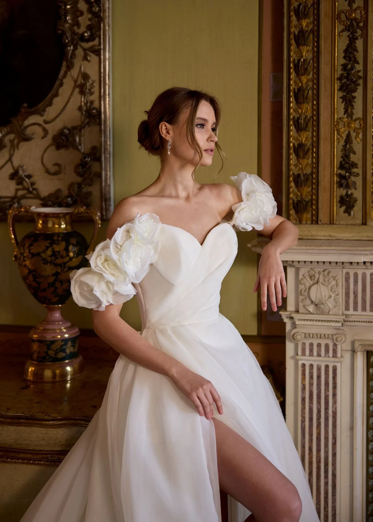 Uma mulher em um elegante vestido branco com mangas florais sopradas, de pé em uma sala decorada com móveis antigos e decoração ornamentada, olhando pensativamente para o lado.