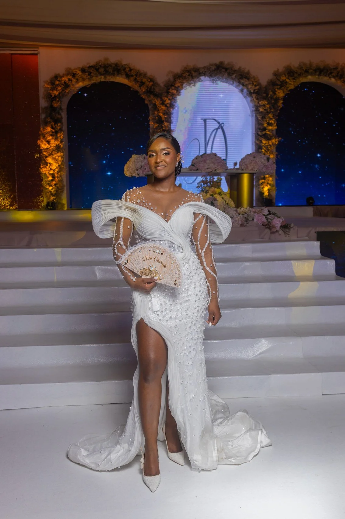 Uma mulher em um vestido branco, embelezado segurando um fã decorativo, de pé em uma escada em um casamento ou evento com flores e estreladas decorações temáticas do céu no fundo.