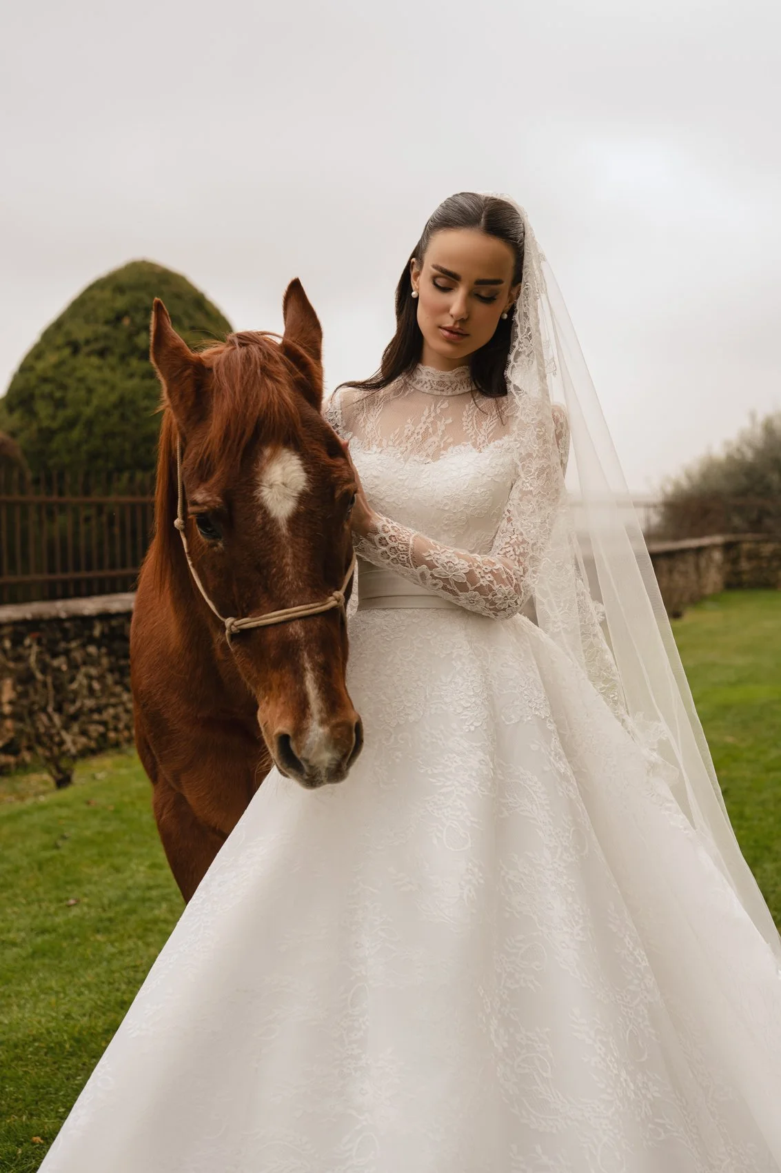 Uma mulher em um vestido de noiva de renda com um véu de pé ao lado de um cavalo marrom com uma marca em forma de estrela branca em sua testa ao ar livre.
