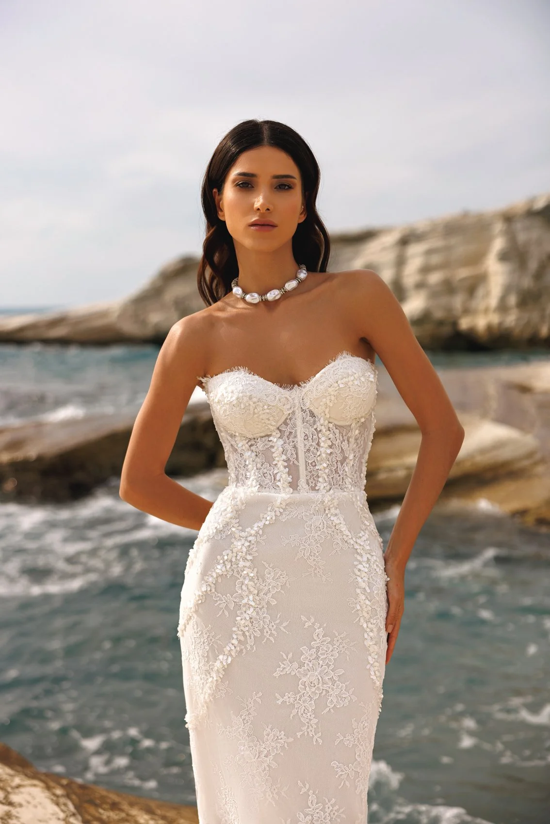 Uma mulher em um vestido de noiva sem alças com detalhes de renda fica em uma costa rochosa ao lado do oceano, vestindo um colar de pérolas e com cabelo escuro e ondulado.