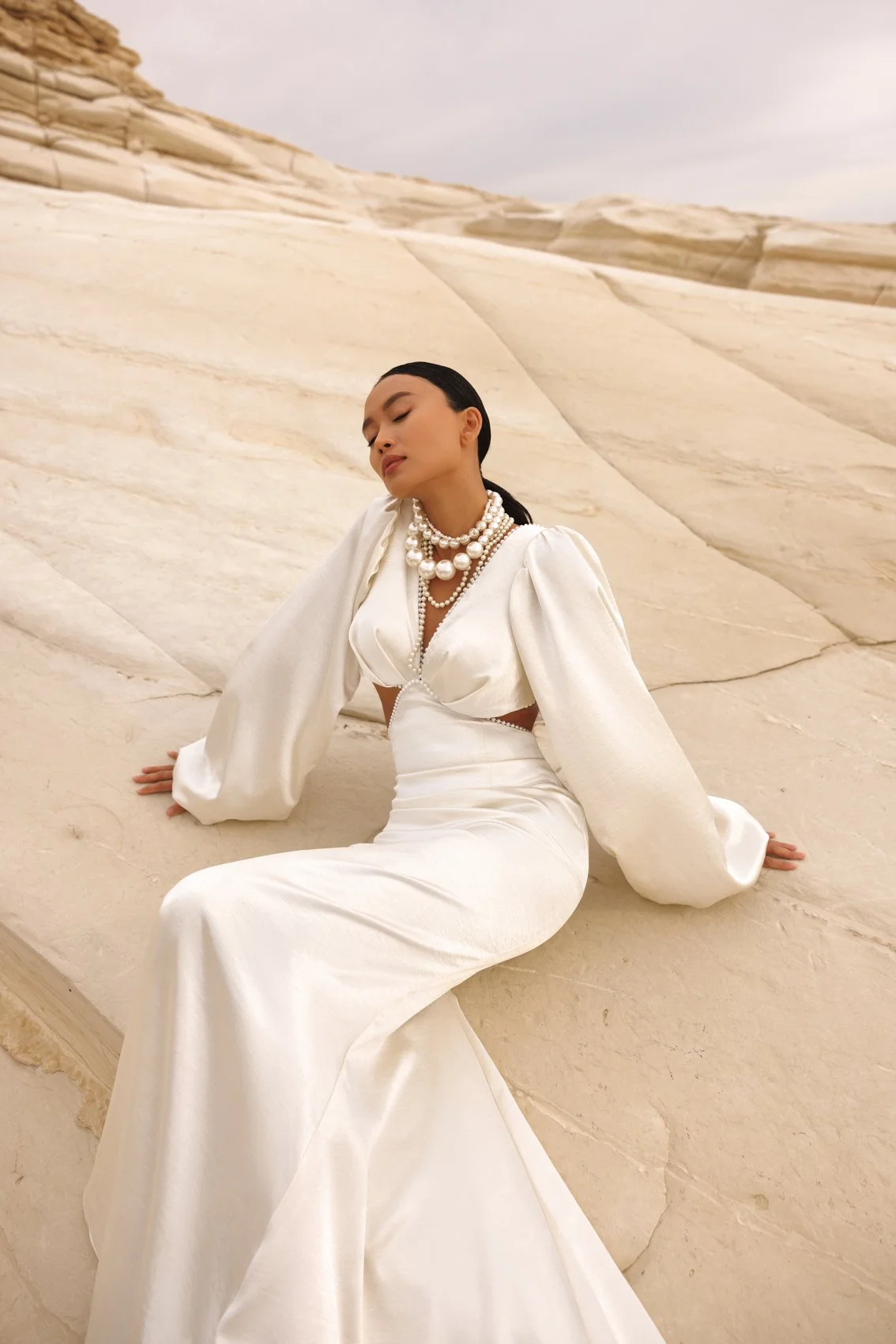Uma mulher vestida em um elegante vestido de cetim branco com mangas folhadas e uma saia enrugada, sentada contra uma paisagem rochosa, cor de creme. Ela usa colares de pérola em camadas, tem cabelo escuro trançado.