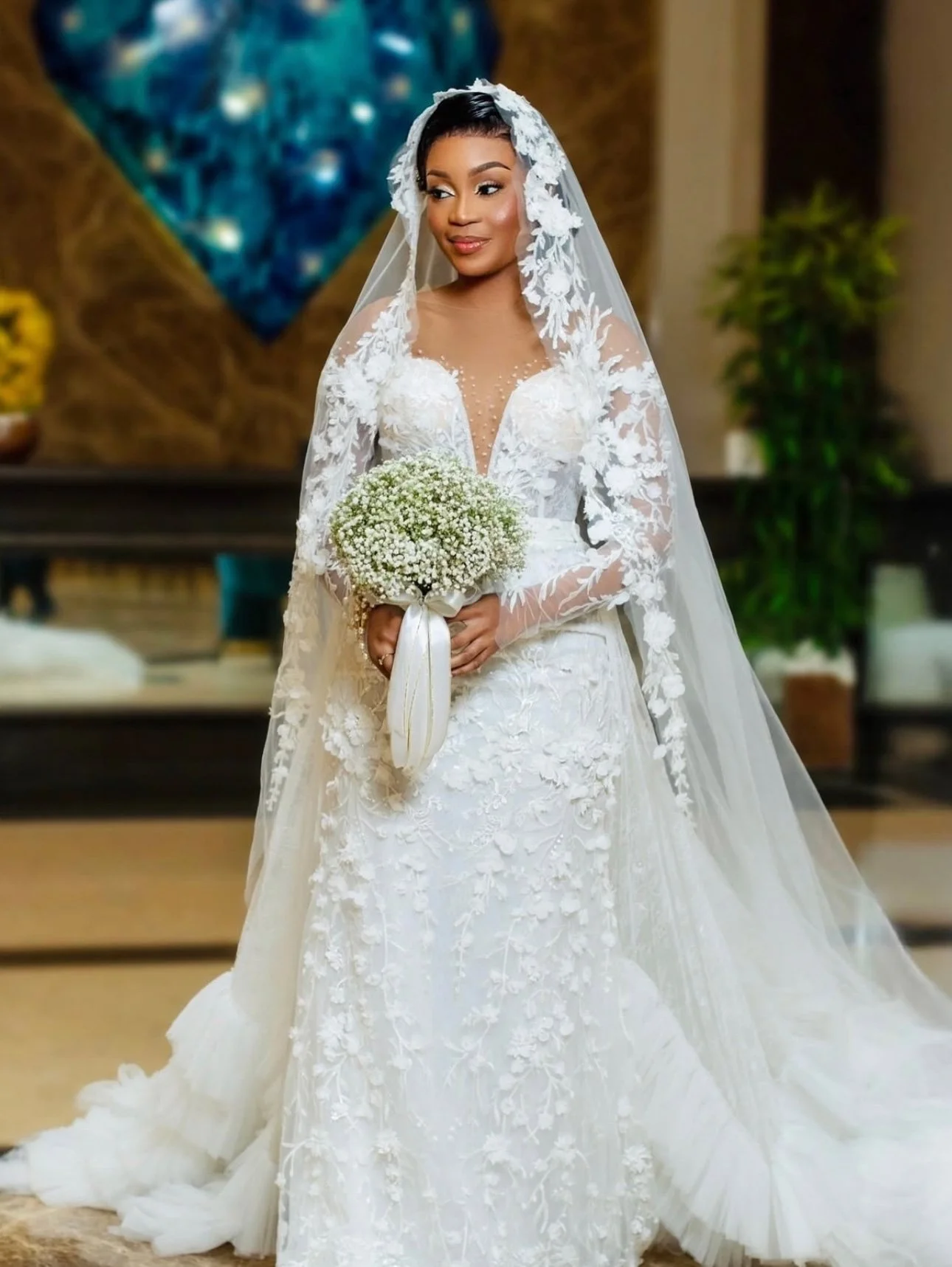Mulher em vestido de noiva branco com véu e buquê de flores pequenas, em ambiente interno decorado.