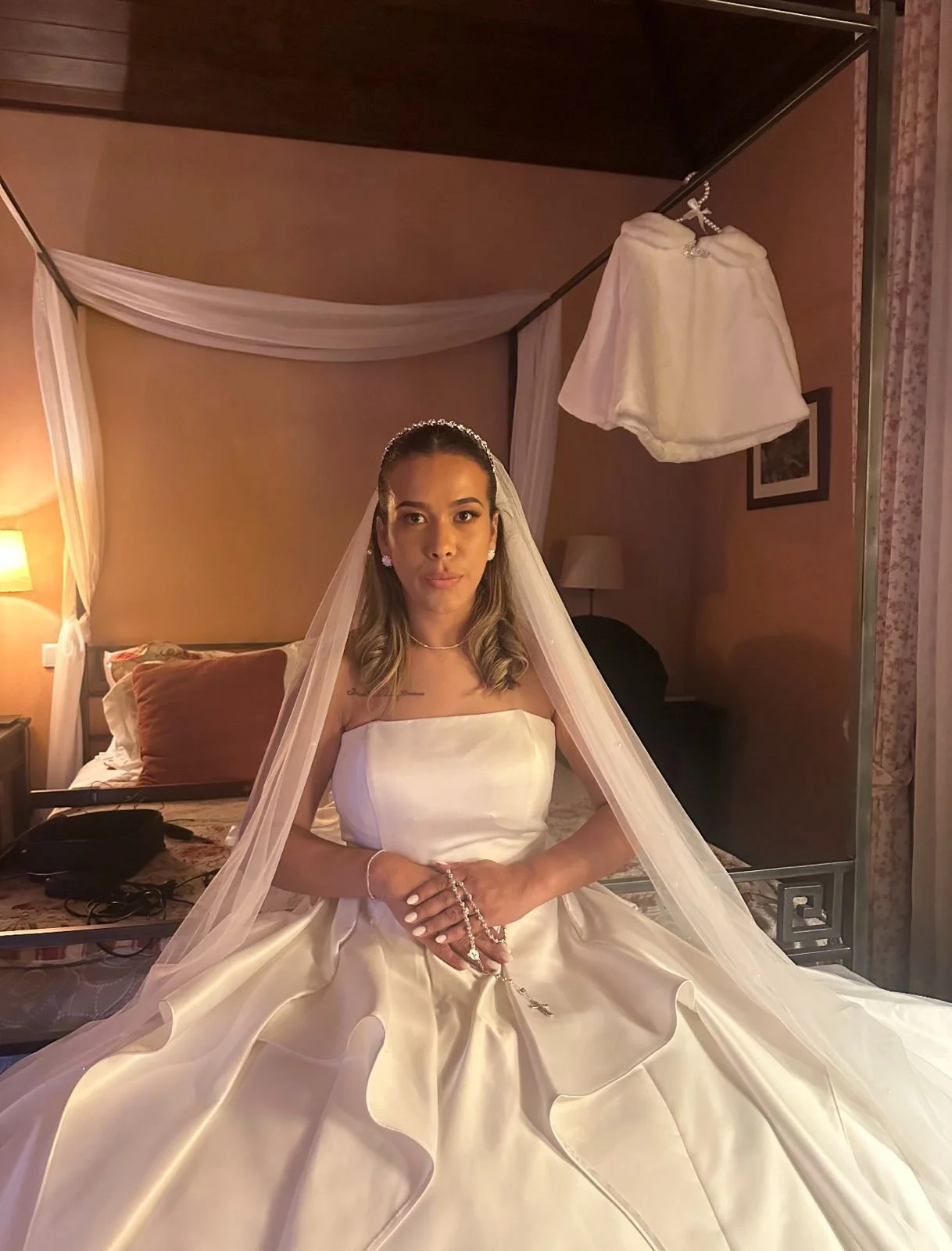 Mulher de vestido de noiva sentada em um quarto, com um véu longo e uma saia de casamento rodada, segurando um rosário. No fundo, uma cama com cabeceira de almofadas, uma lâmpada e uma cortina floral.