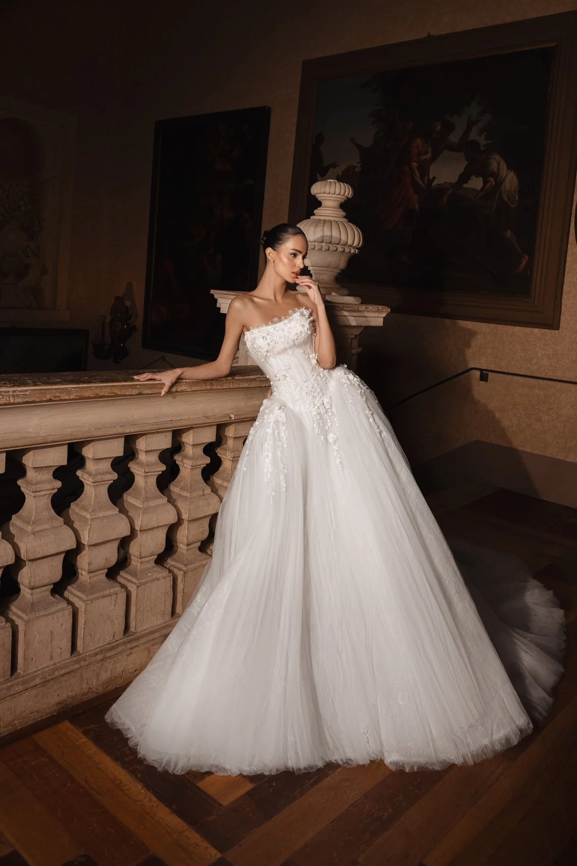 Mulher com vestido de casamento branco em um ambiente com decoração clássica, segurando uma grade de pedra e com um quadro de pintura ao fundo.