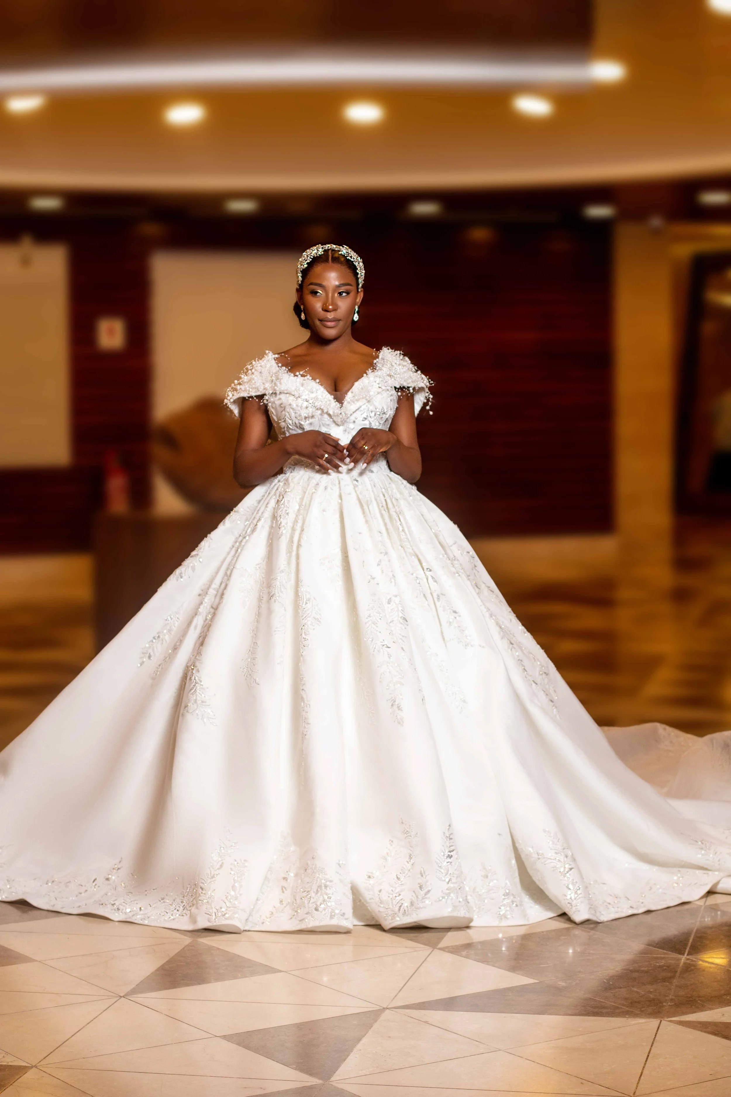 Mulher deslumbrante em vestido de casamento branco com detalhes bordados, usando tiara e brincos, posando em ambiente indoor com piso de ladrilhos decorativos.