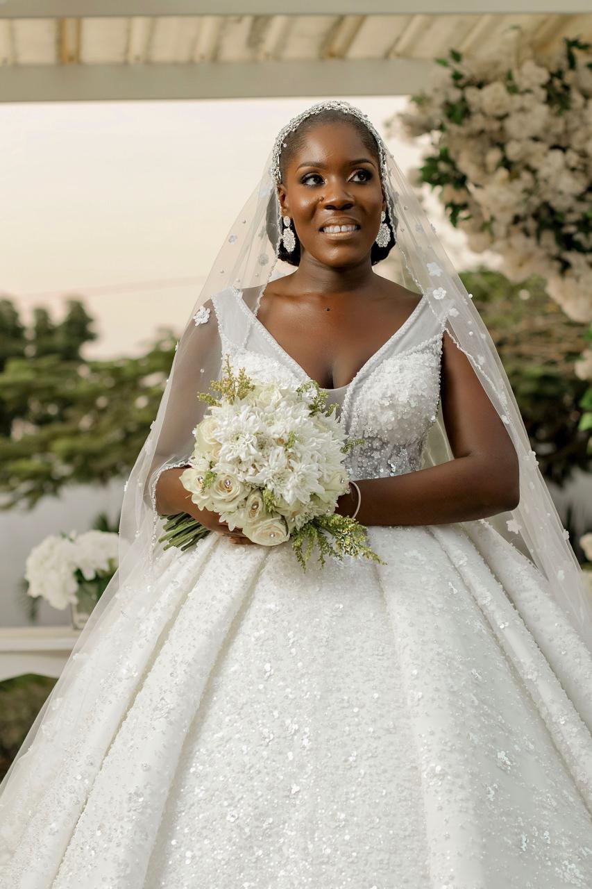 Noiva com vestido de casamento branco e véu, segurando um buquê de flores brancas, em um cenário externo.