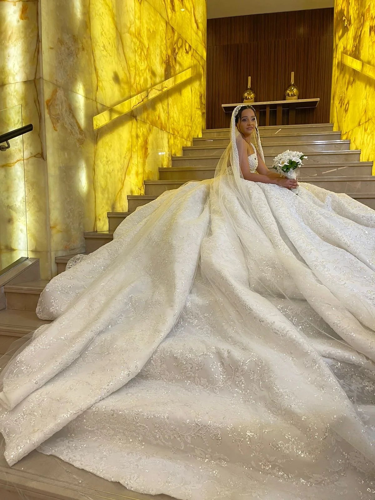 No centro da imagem, uma mulher vestida com um vestido de casamento branco, com longo véu e segurando um buquê de flores brancas. Ela está sentada em uma escadaria de mármore, com paredes de mármore dourado ao fundo, decoradas com vasos dourados.