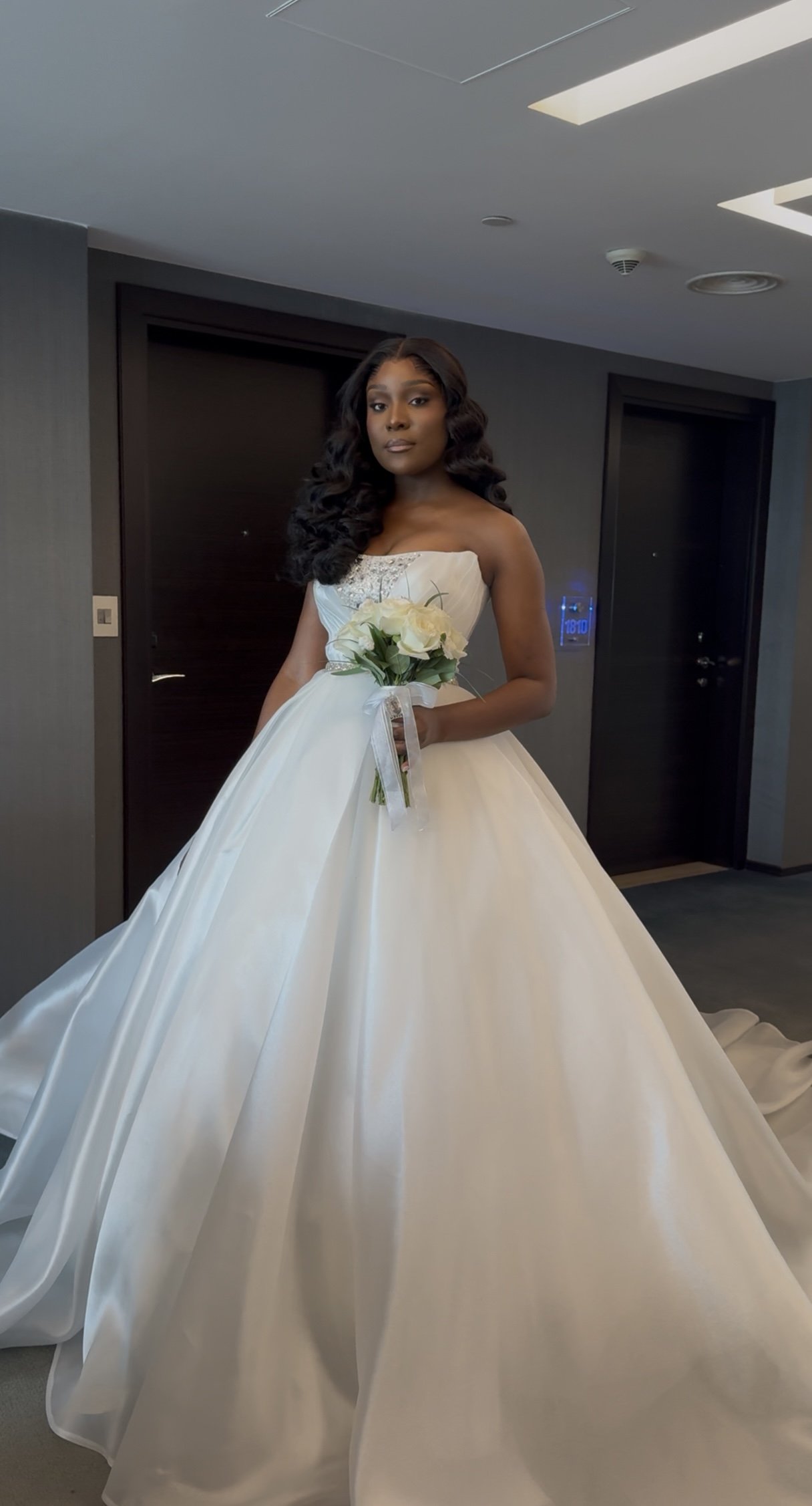 Mulher com vestido de noiva, penteado cacheado, segurando buquê de flores brancas em um ambiente de hotel ou salão.