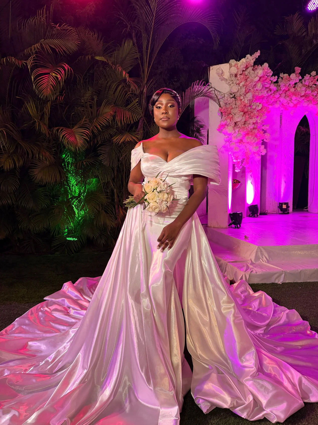 Uma mulher em um vestido de casamento branco segurando um buquê de rosas em um local de casamento decorado com iluminação rosa e roxo, vegetação e arranjos florais.