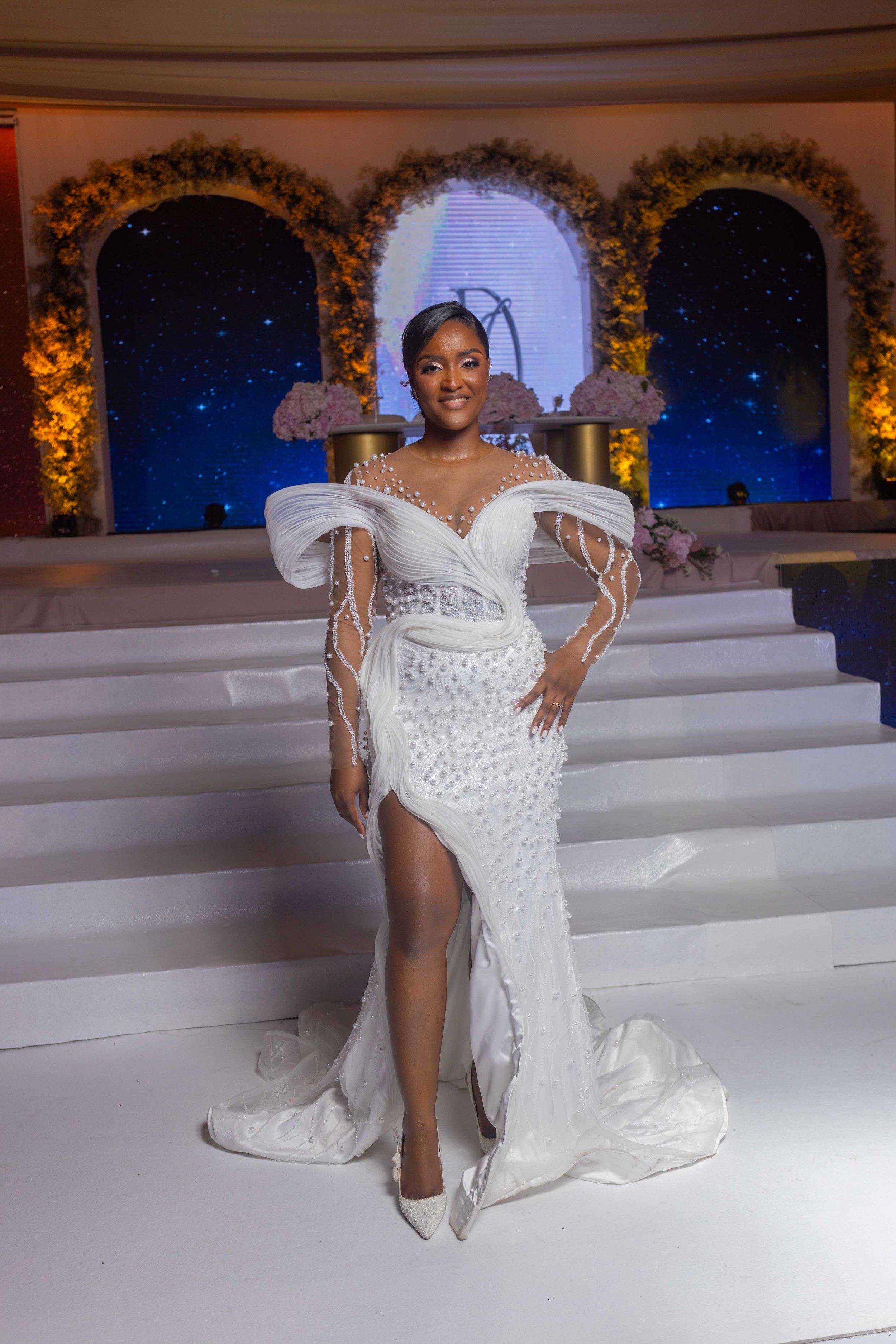 Mulher elegante em vestido de casamento branco com detalhes de pérolas, sorrindo, em escada de palco decorada com flores e fundo de espaço estrelado.
