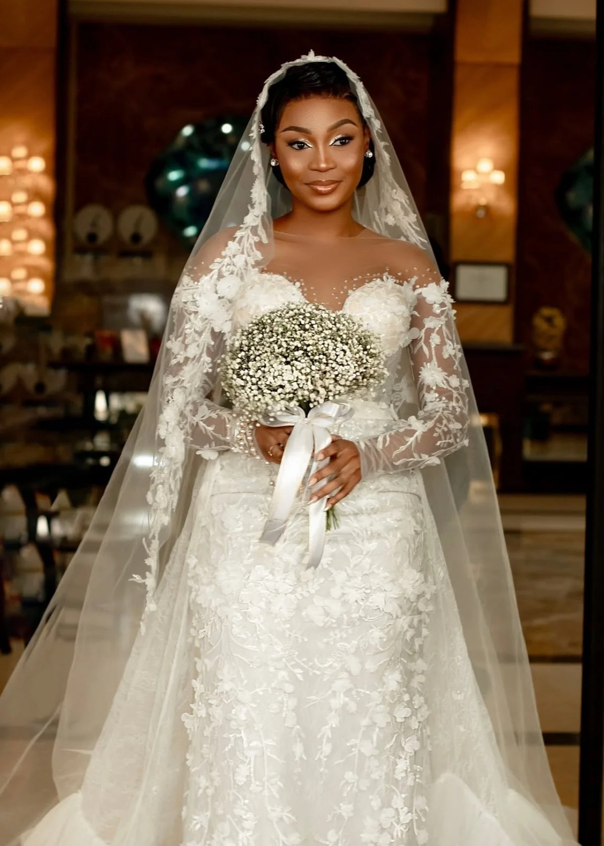 Mulher afro-brasileira vestida de noiva com vestido branco de renda, véu e buquê de rosas brancas, em ambiente interno decorado com objetos de decoração e iluminação quente.