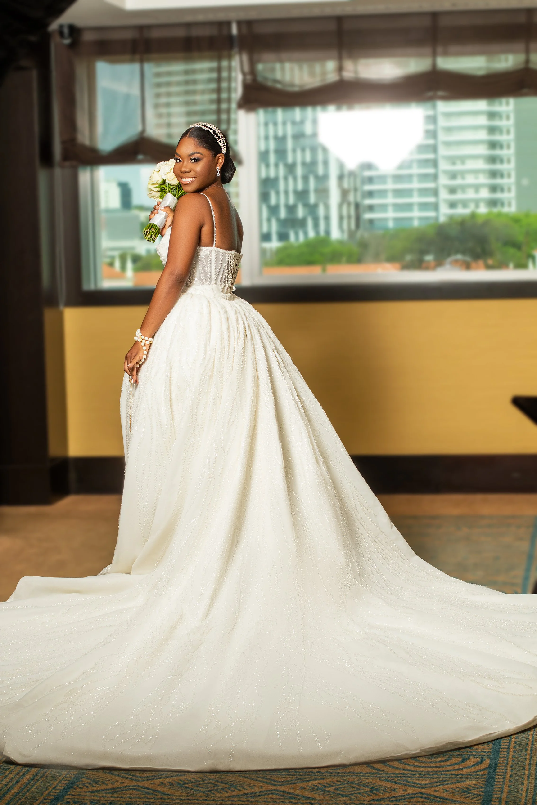 Mulher sorridente em um vestido de noiva branco, segurando um buquê de flores, dentro de um ambiente com janelas de vidro e prédios ao fundo.
