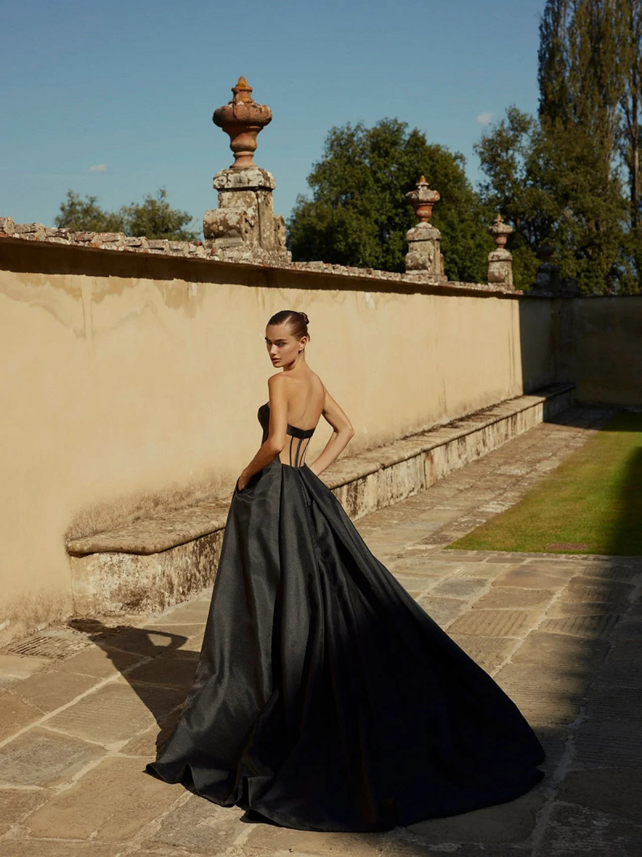 Uma mulher em um vestido preto sem alças com um longo trem fica ao ar livre por uma parede de pedra com urnas no topo, sob um céu azul claro cercado por árvores.