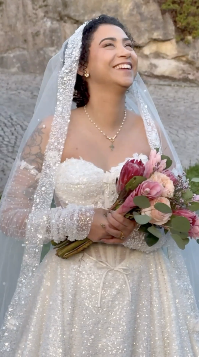 Uma noiva sorridente com cabelo encaracolado escuro, vestindo um vestido de noiva branco com detalhes frisados, segurando um buquê de flores cor-de-rosa e vermelhas, em pé ao ar livre com terreno rochoso no fundo.