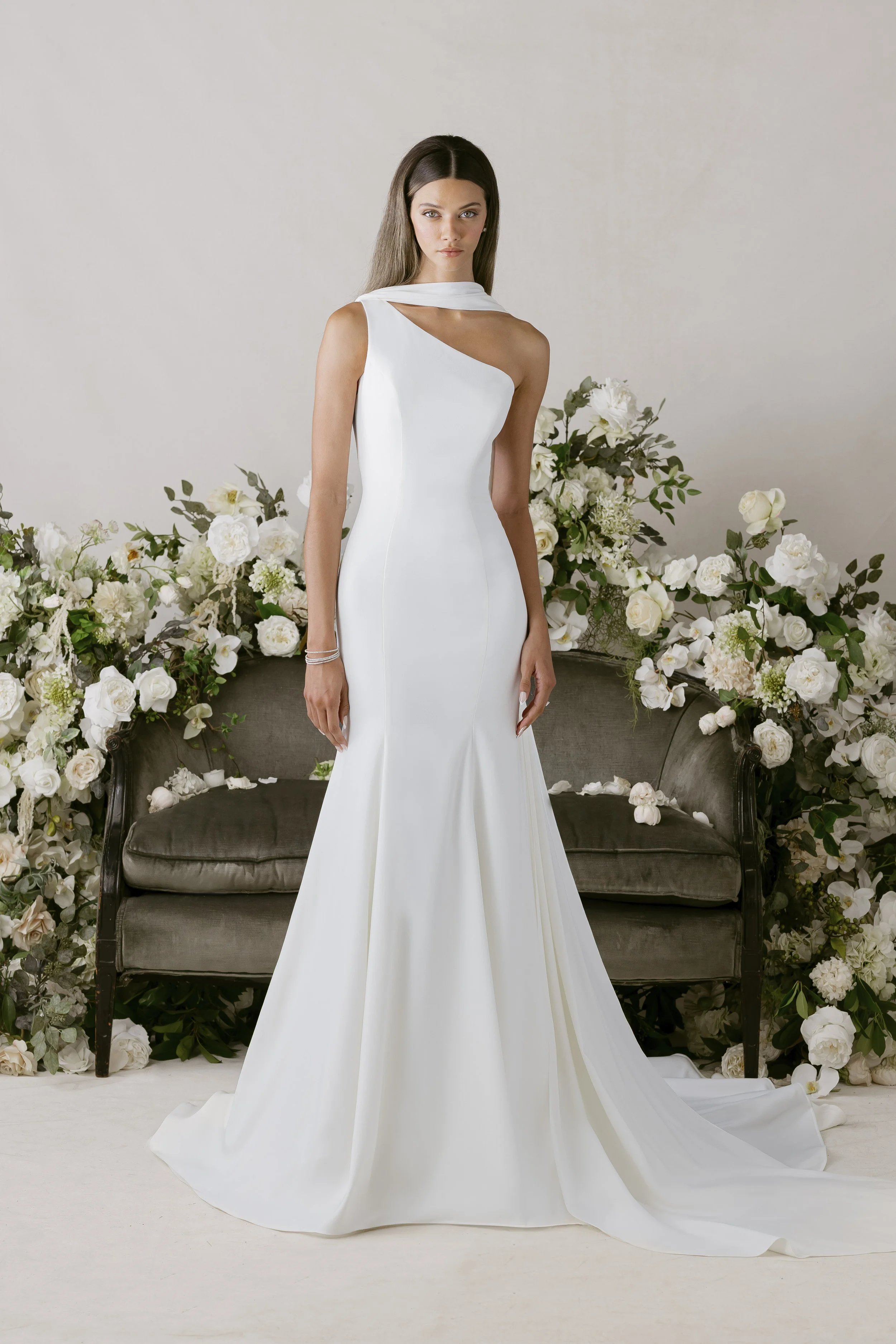 Mulher vestida com um vestido de casamento branco longo e ajustado, com um arranjo de flores brancas ao fundo e um sofá antigo de cor escura.