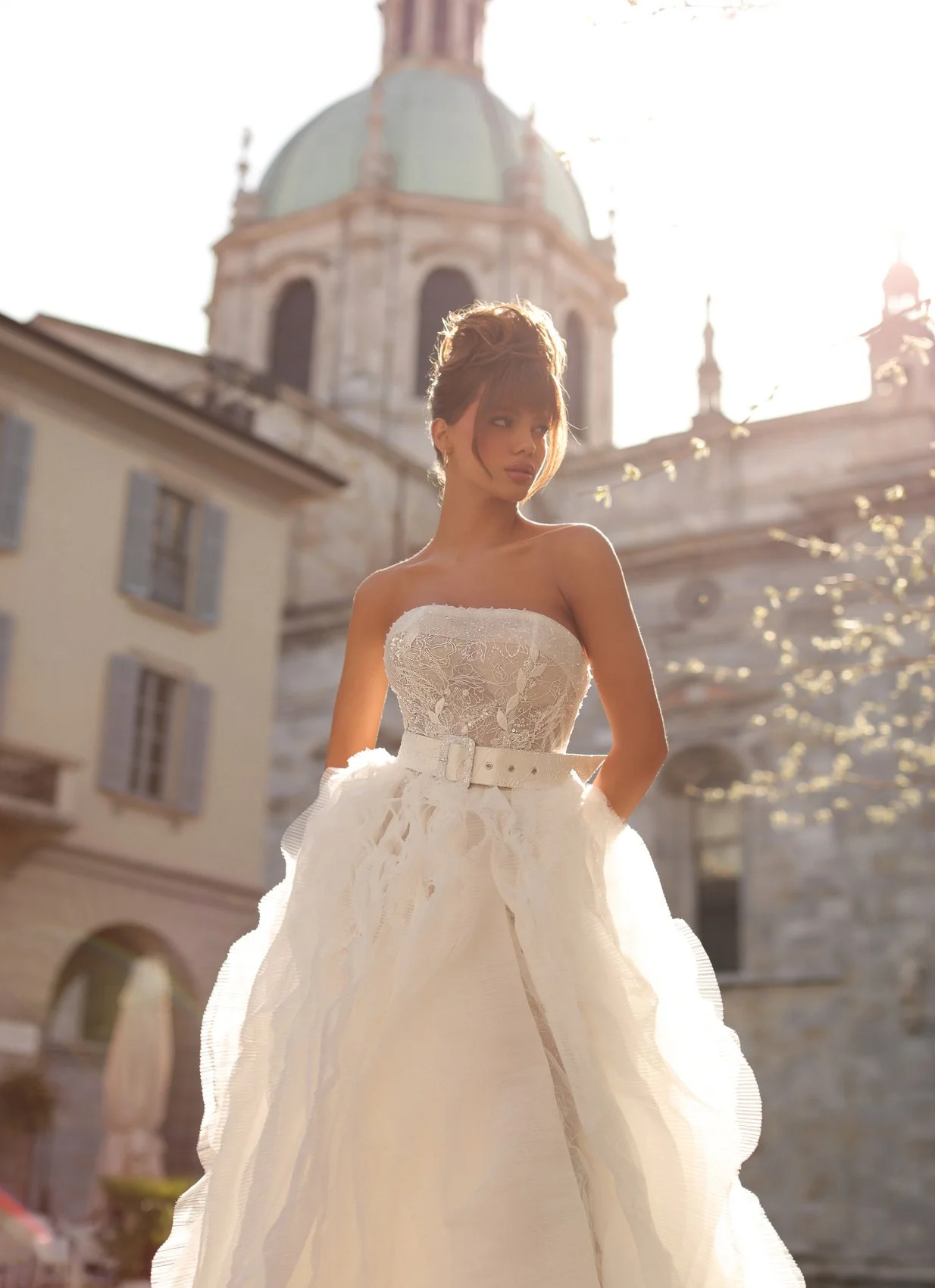 Uma mulher em um vestido de noiva branco sem alças com detalhes de renda e um cinto, de pé ao ar livre na frente de um edifício histórico com uma cúpula, durante a luz solar brilhante.