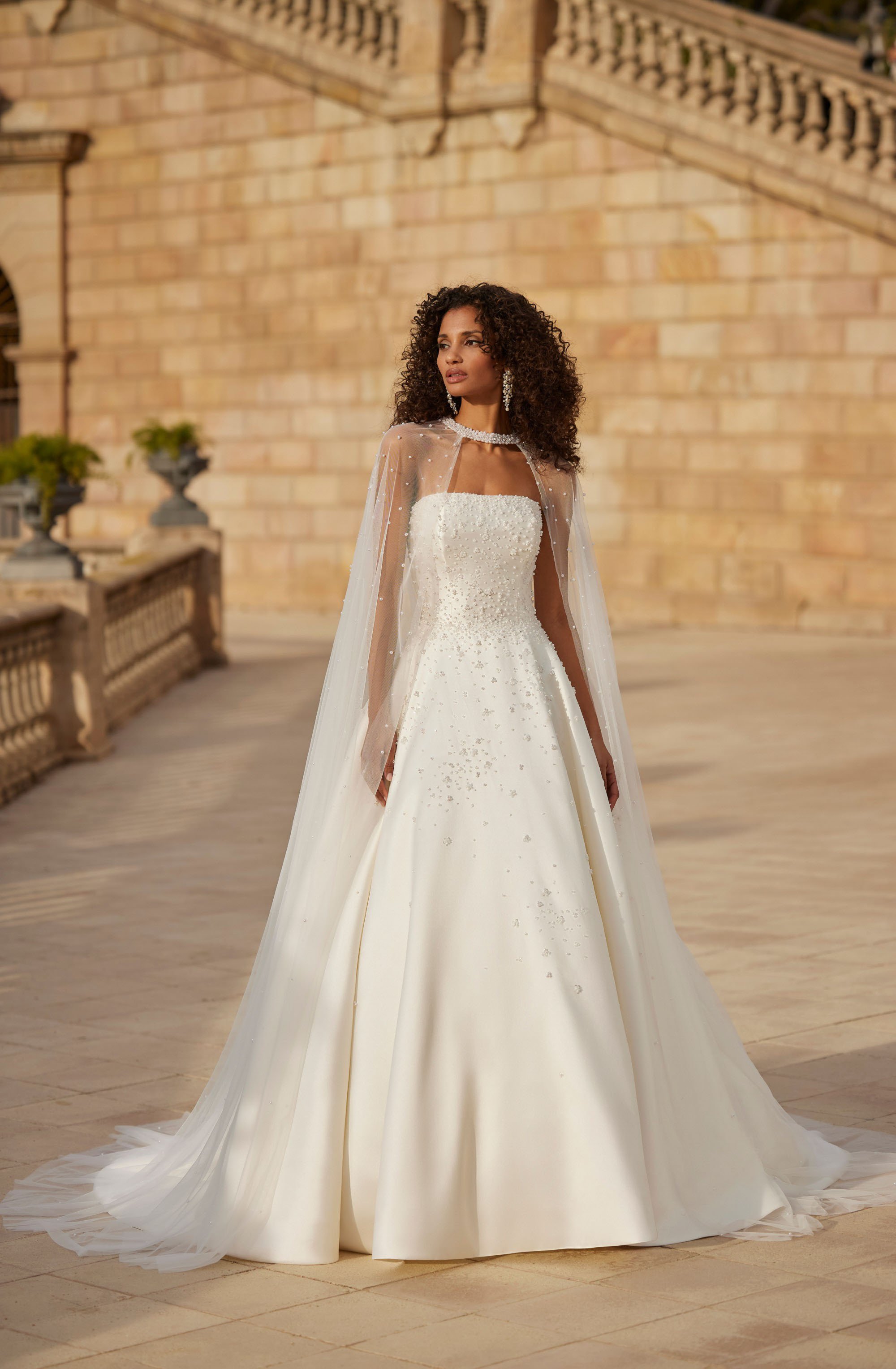 Noiva vistindo vestido de casamento branco, com véu longo, de costas, em um fundo de cortina azul. O vestido tem uma cauda com detalhes em forma de folha no chão.