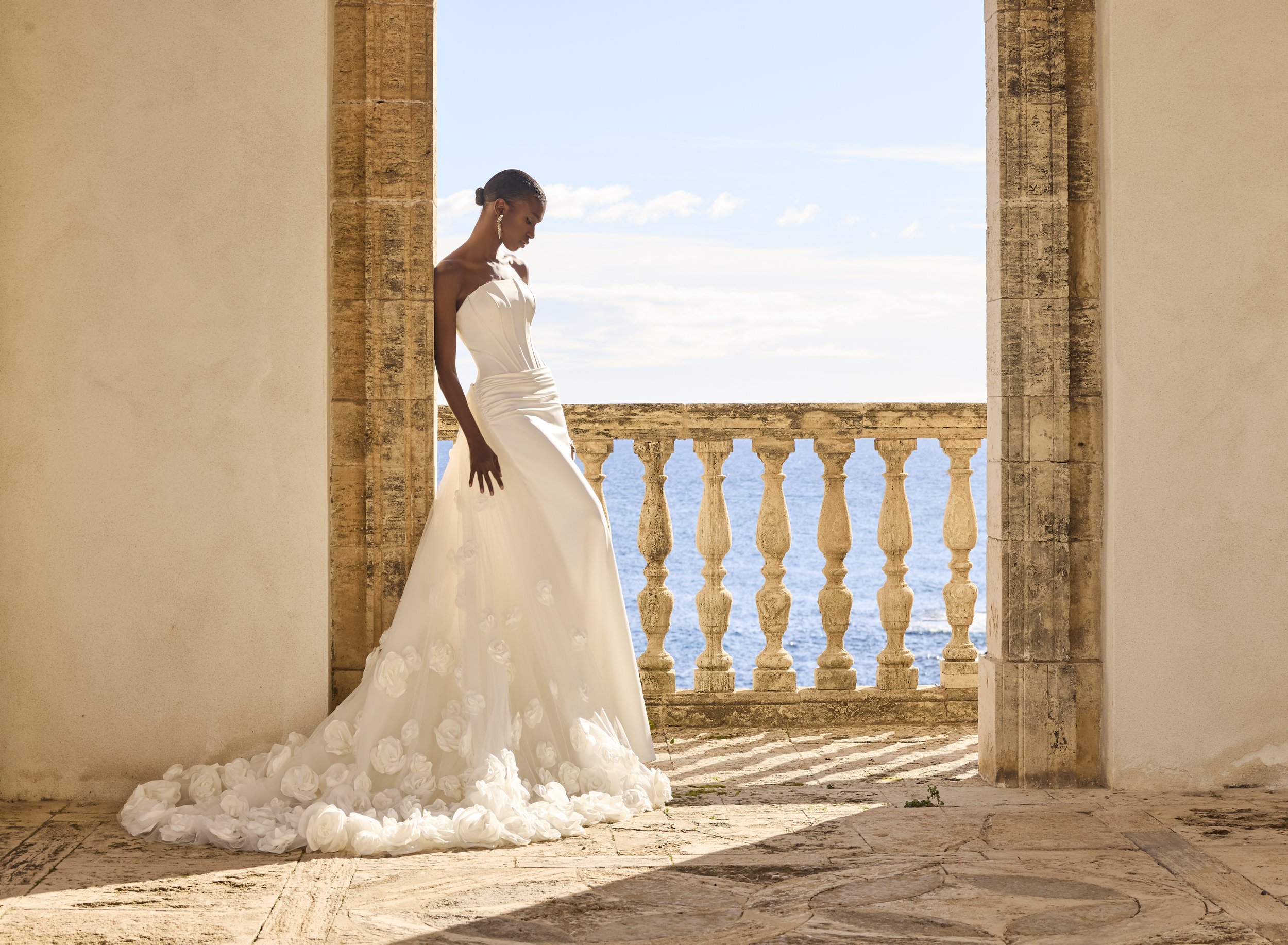 Mulher vestida com vestido de noiva branco, de cabelos presos, na varanda com vista para o mar e céu nublado.