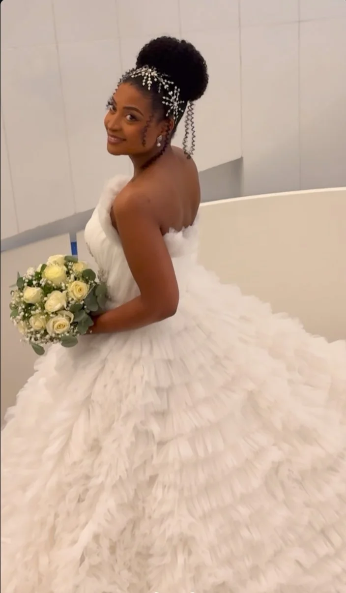Uma noiva em um vestido de casamento branco sem alças com folhos em camadas, segurando um buquê de rosas brancas e vegetação, com o cabelo estilizado adornado com pérola e acessórios de cabelo de cristal em um coque alto.