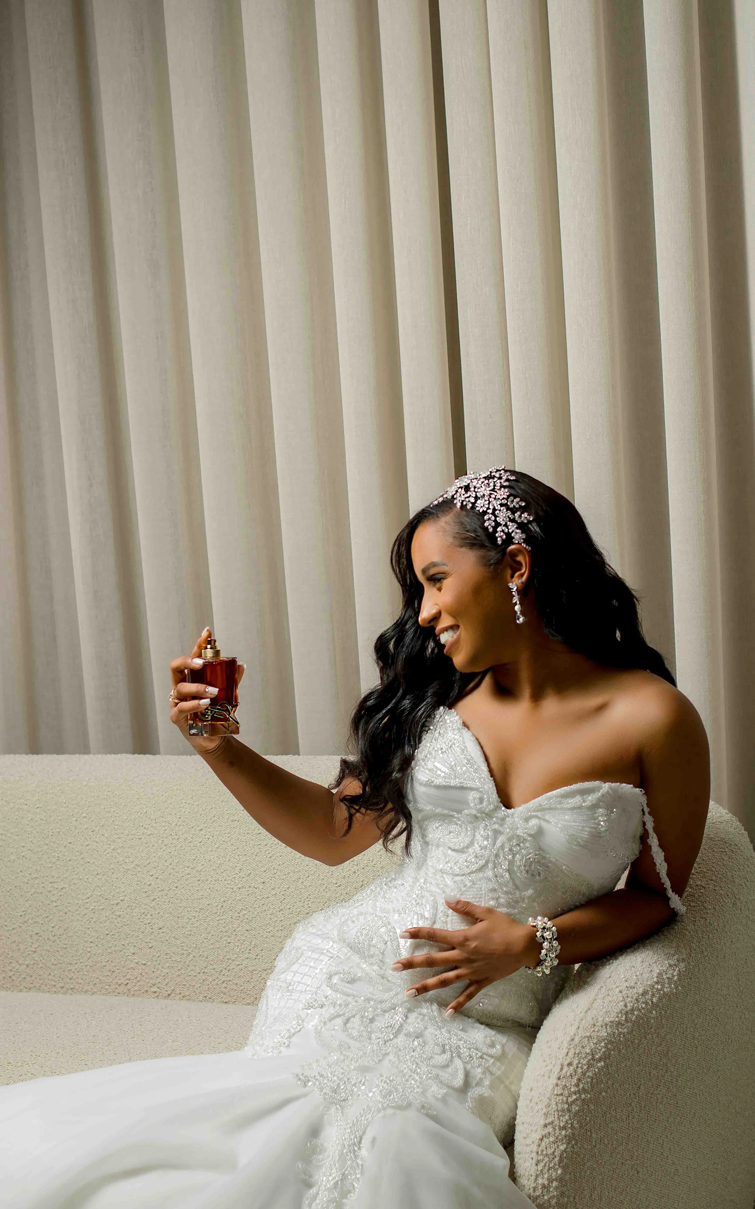 Mulher vestida de casamento sentada em sofá, segurando perfume e sorrindo, com fundo de cortinas.