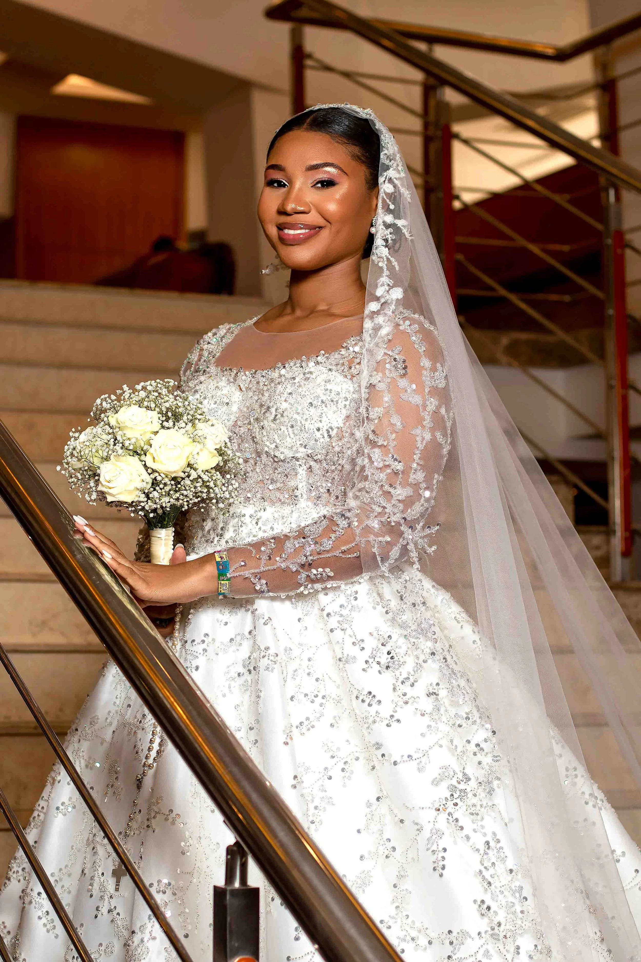 Uma noiva sorridente vestindo um vestido de casamento branco com detalhes brilhantes, segurando um buquê de rosas brancas e usando um véu de renda. Ela está em um ambiente interno com escadas ao fundo.