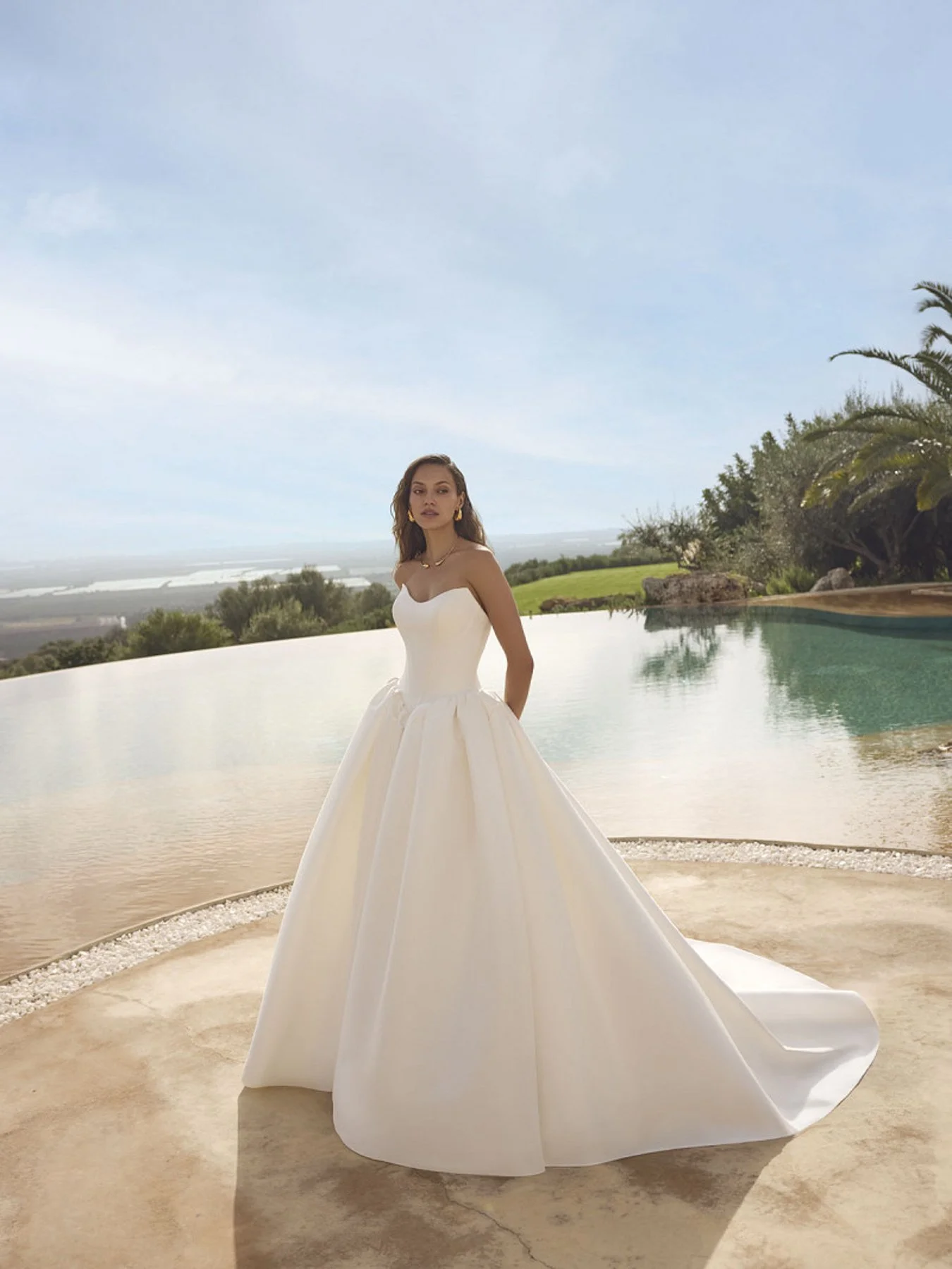 Mulher usando vestido de casamento branco ao lado de piscina, com paisagem natural ao fundo.