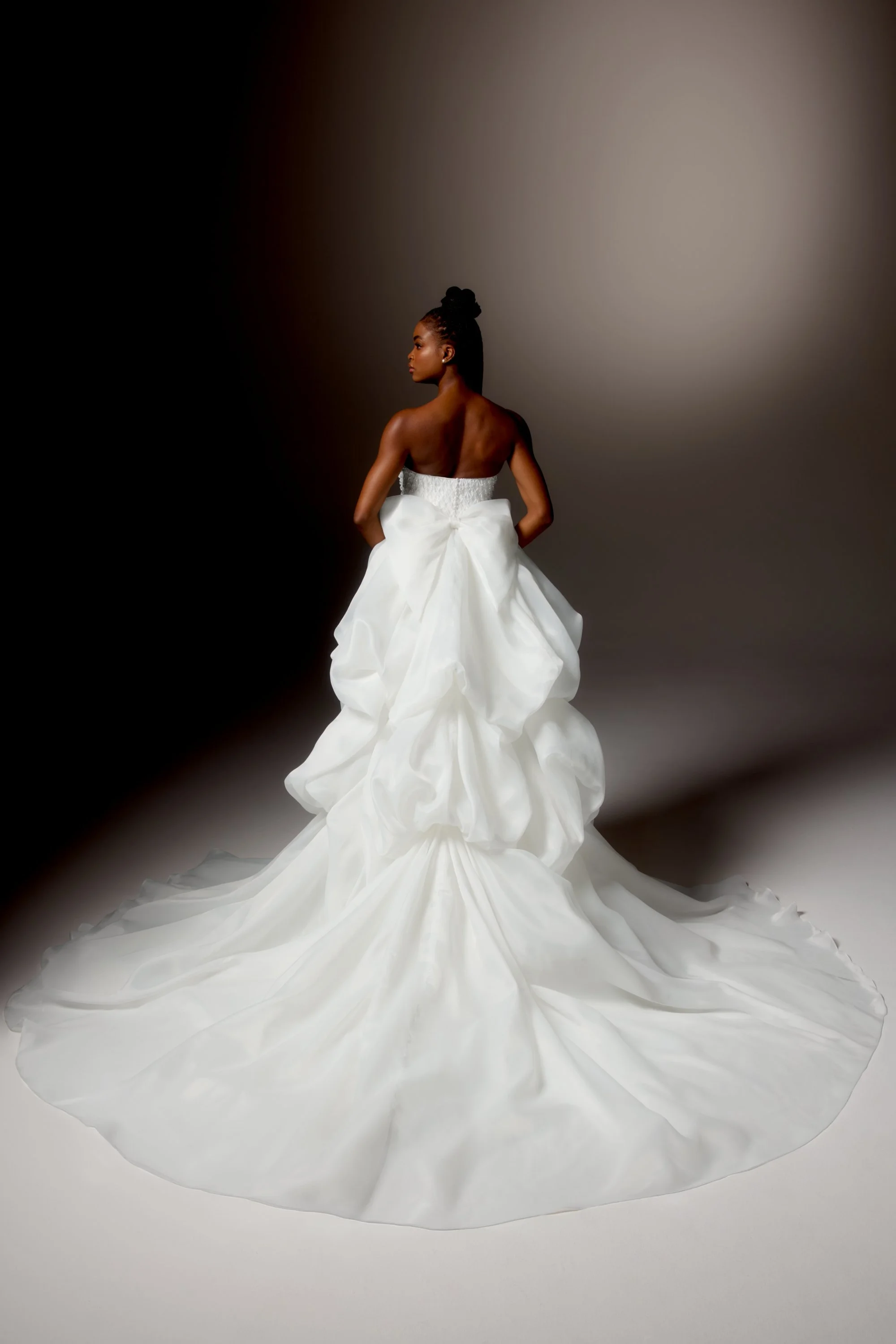 Mulher jovem de pele escura usando vestido de noiva branco com saia volumosa e corpete ajustado, posando de lado em um fundo cinza escuro.