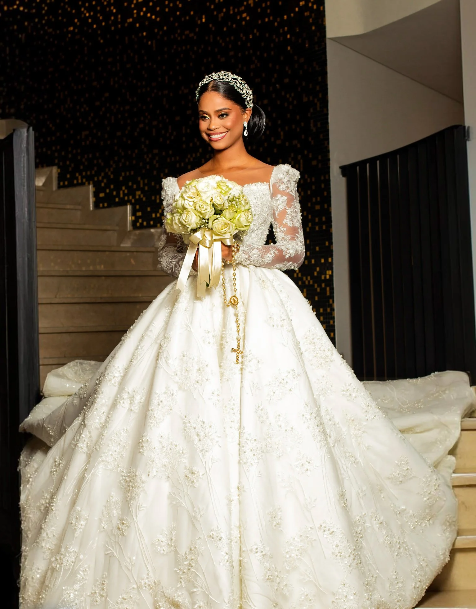 Noiva sorridente com vestido de casamento branco com detalhes de renda e bordados, usando tiara de cristais e brincos, segurando buquê de rosas brancas com fita dourada, em ambiente interno.