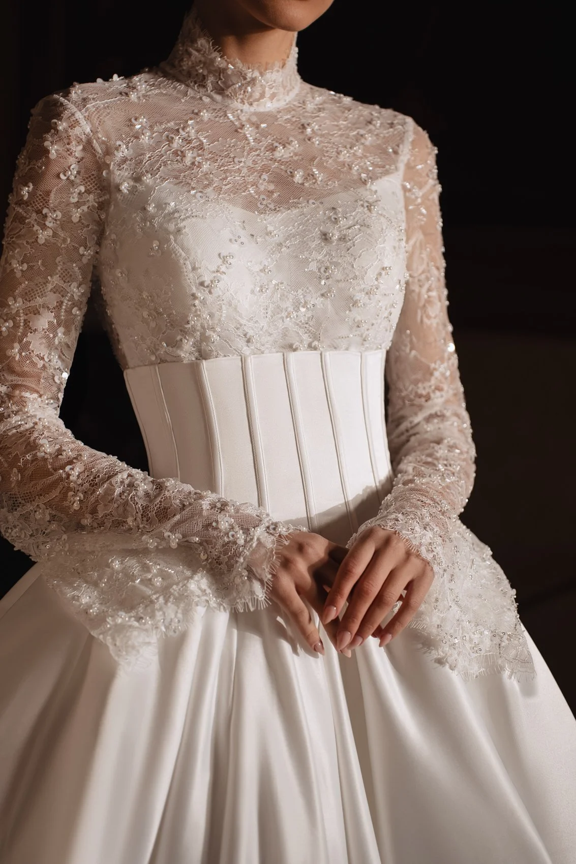 Close-up de uma mulher usando um vestido de noiva branco detalhado com enfeites de renda e pérola, com mangas compridas de renda, decote alto e cintura ajustada.