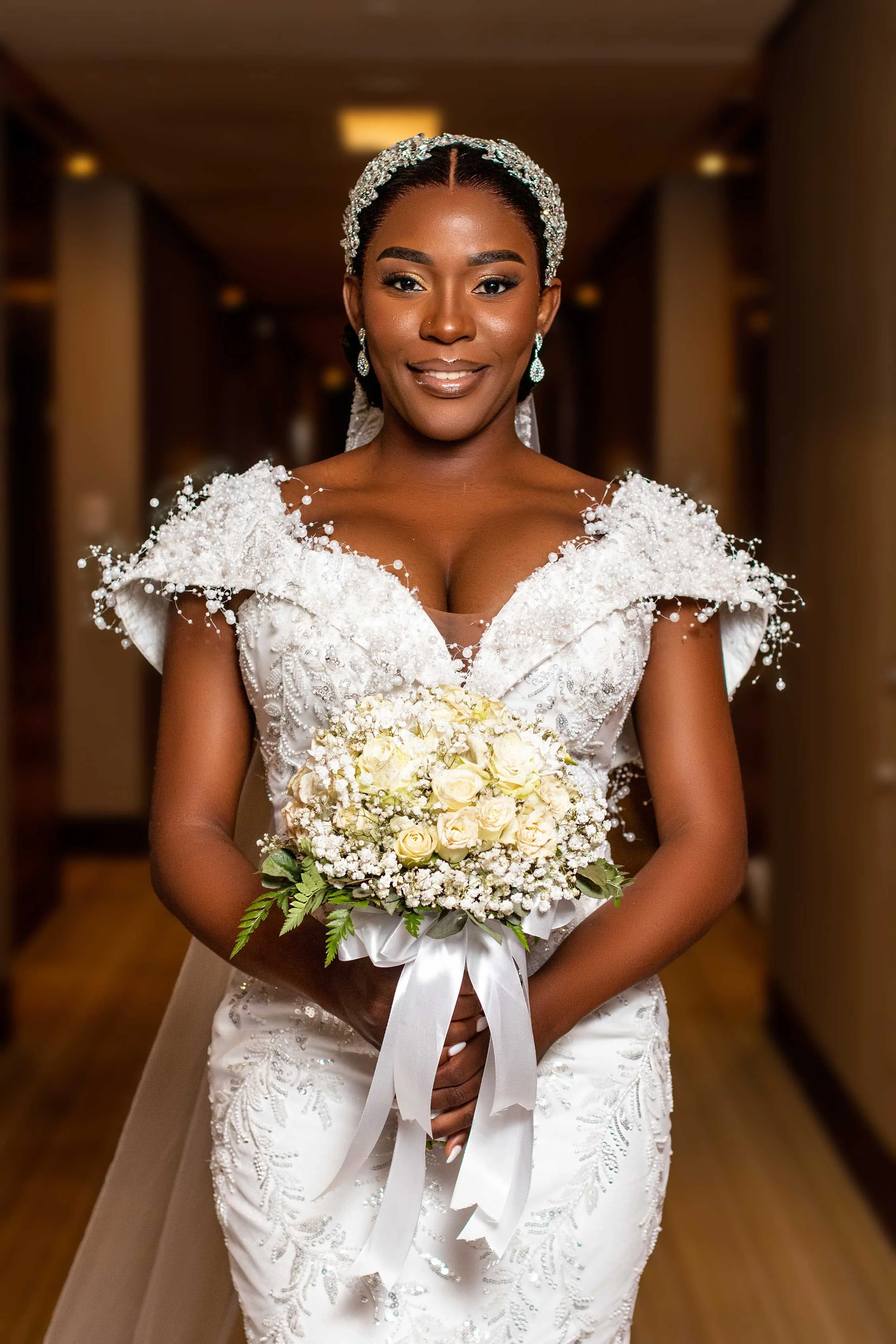 Mulher sorridente, vestida de noiva com vestido branco com detalhes em pérolas, usando tiara de pedras brilhantes, brincos e maquiagem, segurando um buquê de flores brancas, em ambiente interno com iluminação quente.