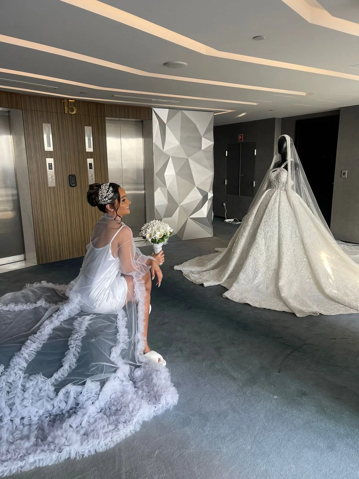 No interior de um prédio, uma mulher com vestido de noiva revela um vestido de casamento elegante em cima de um manequim. Ela está com um vestido de cetim branco com renda e uma tiara, segurando um buquê de flores. A mulher está sentada no chão, sorr