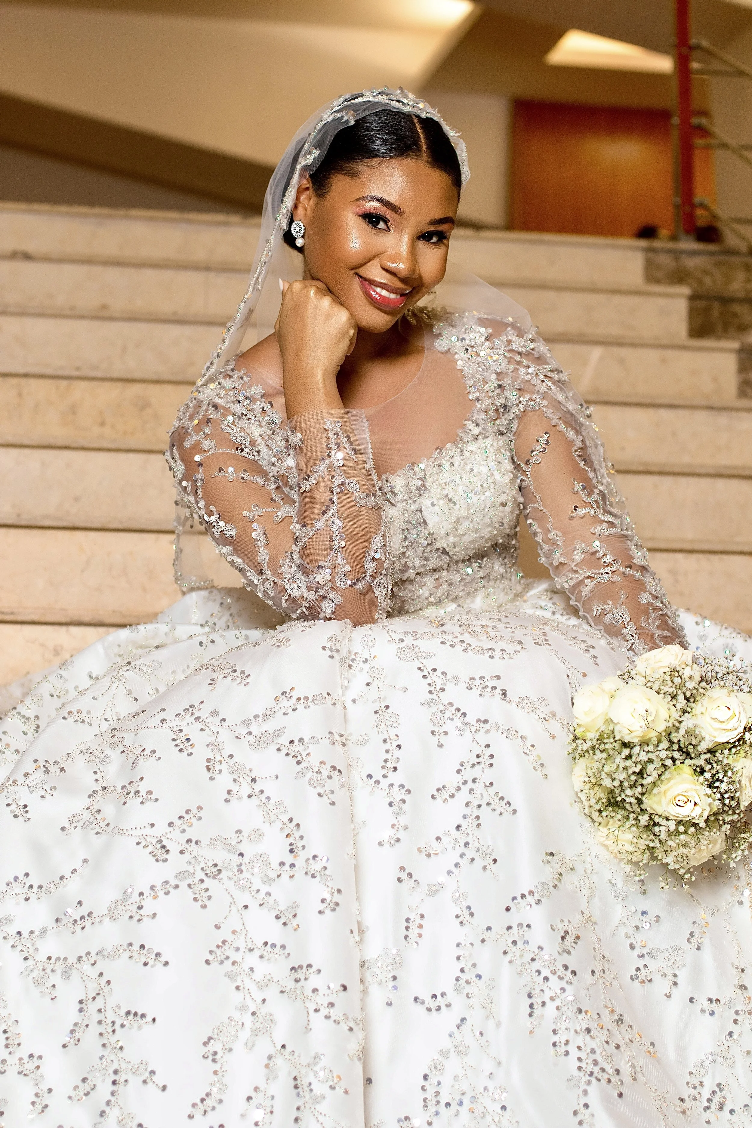 Mulher sorridente vestida com vestido de casamento branco com detalhes em pérolas e pedras, usando véu e brincos de diamante, segurando buquê de rosas brancas, em uma escadaria de mármore.