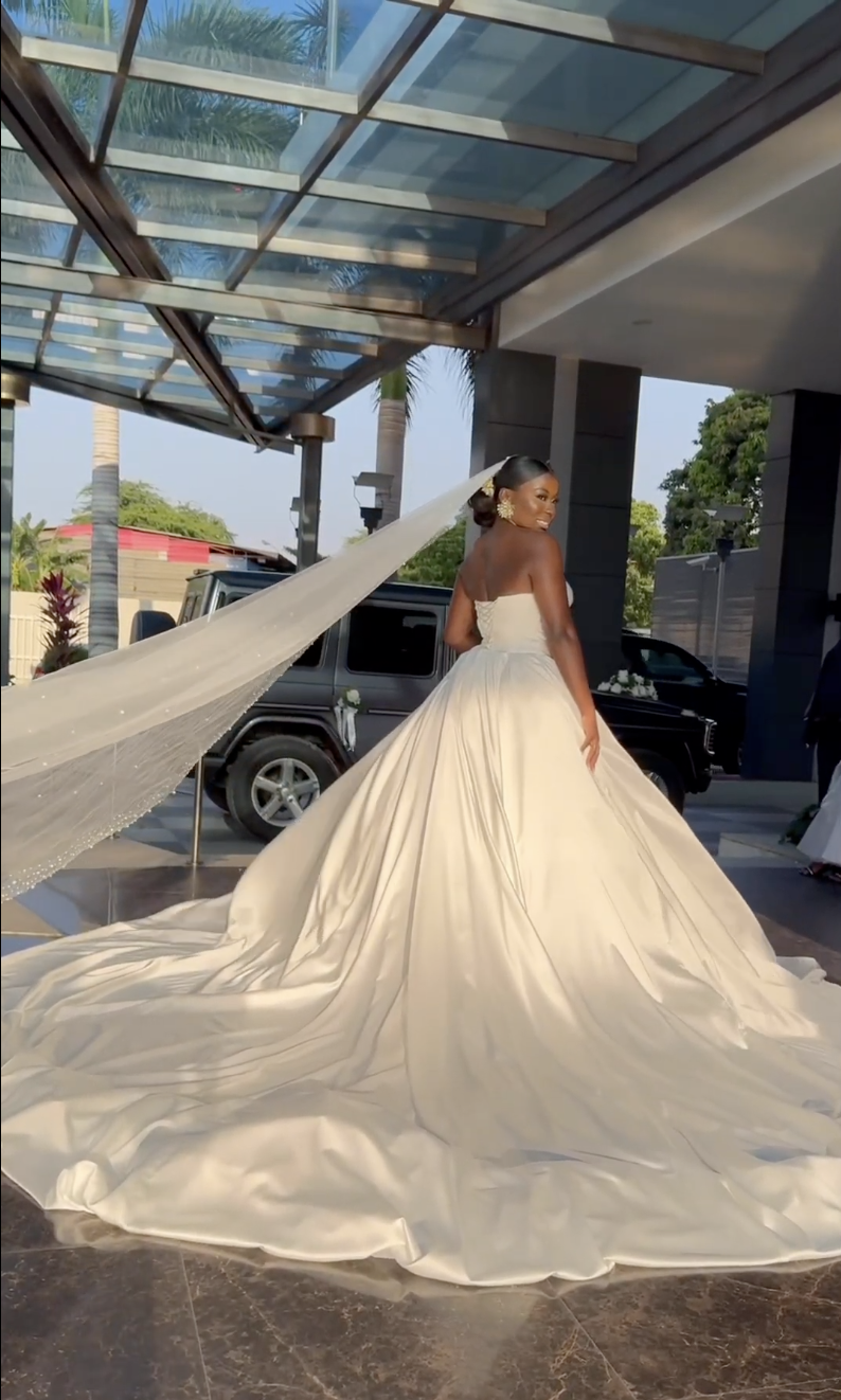 Uma noiva em um vestido de casamento branco sem alças com um trem longo, sob um telhado de vidro em um local ao ar livre, com palmeiras e um veículo preto no fundo.