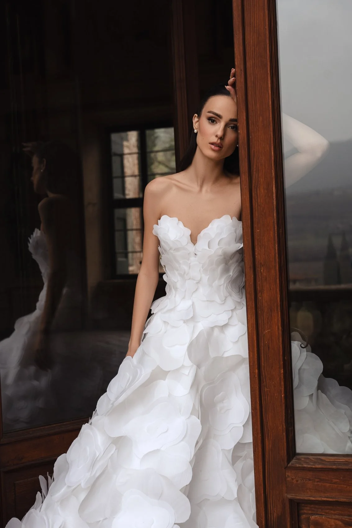 Uma mulher em um vestido de noiva branco sem alças com detalhes florais em camadas, de pé atrás de uma moldura de porta de madeira e olhando para a câmera.