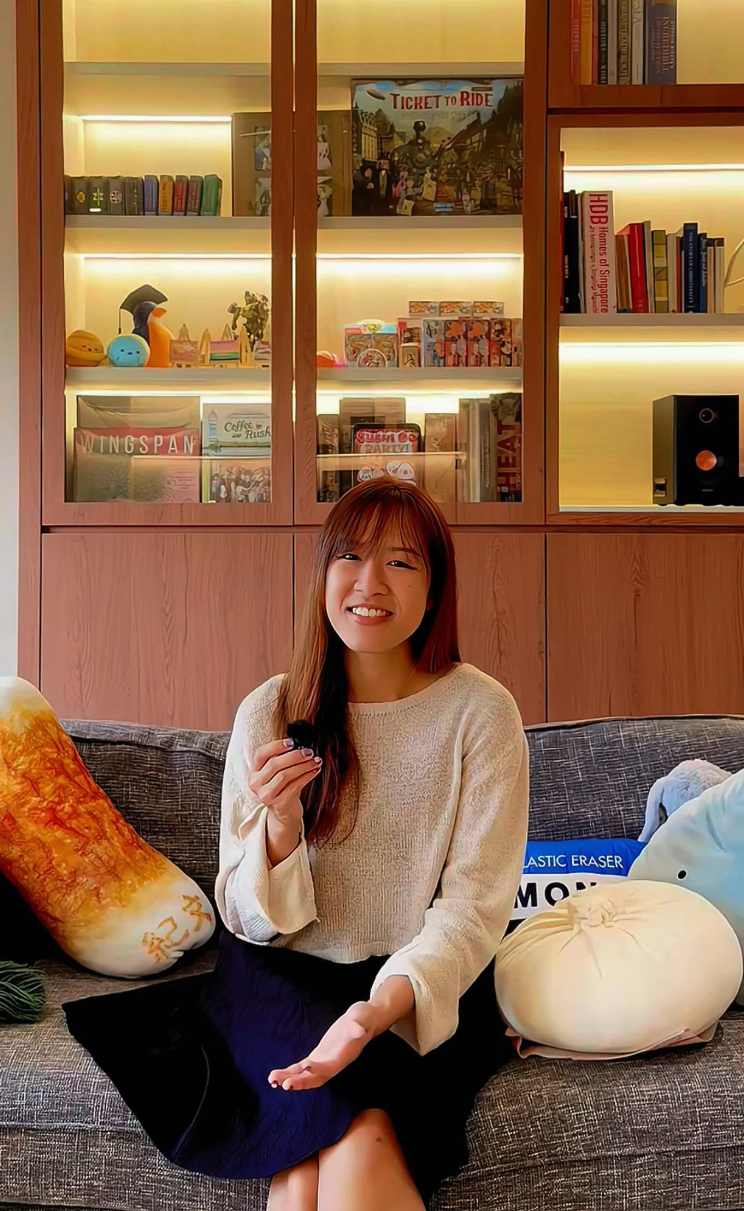 A woman with long brown hair, wearing a beige sweater and black skirt, sitting on a gray couch in a room with a wooden bookshelf. She is smiling, holding a small black object, with decorative pillows on the couch, and various items and books in the background.