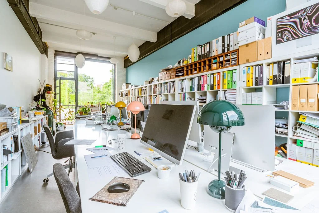 An office with a long white desk, computers, lamps, chairs, and extensive shelving with binders, folders, and paper.