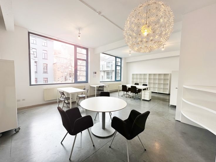 Modern office space with white walls, large windows, and minimalistic furniture including black and white chairs and desks. A round table with four black chairs is in the foreground, and a decorative pendant light hangs from the ceiling.