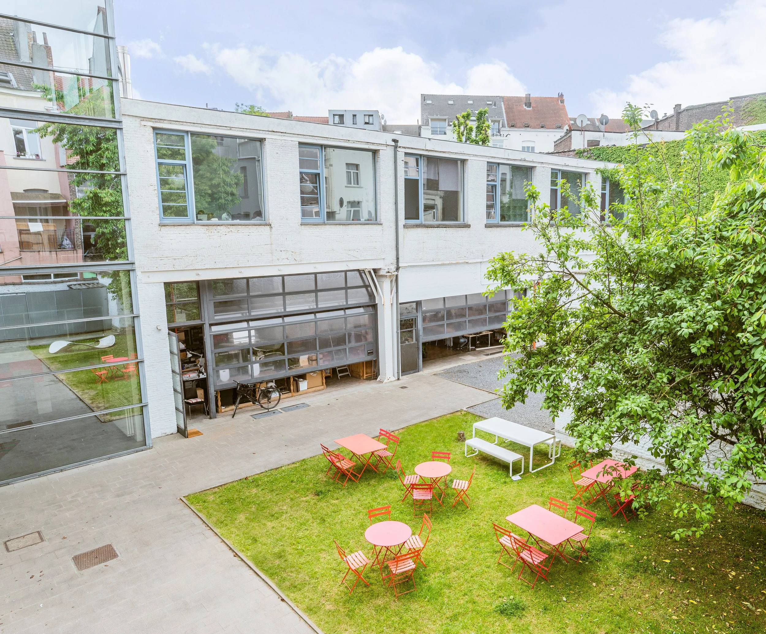 Outdoor scene of a modern building with large glass windows and a small grassy area with pink tables and chairs, also features a white bench, a large leafy tree, and a bicycle near the building.