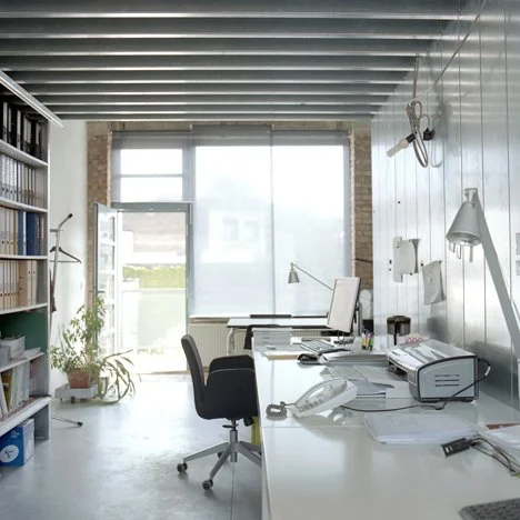 An office workspace with a desk, computer, office chairs, and filing shelves near a large window with sunlight.