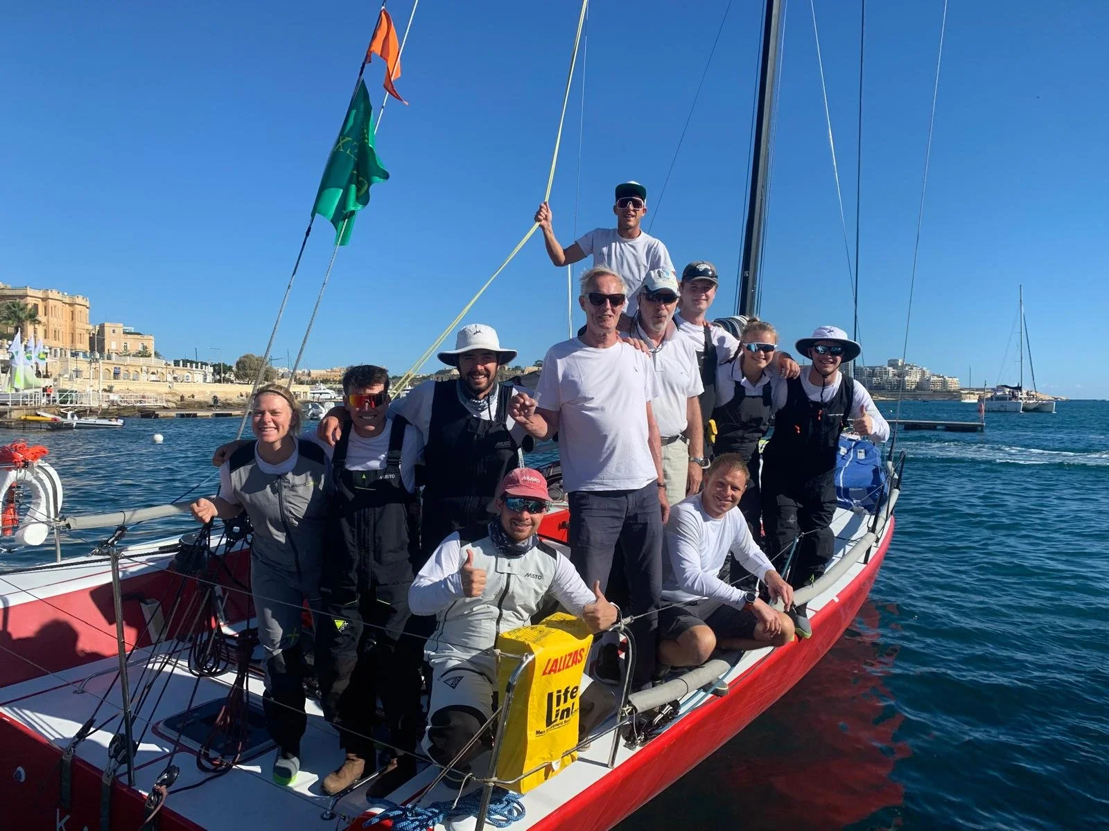 Group of people on a sailing boat in a marina on a sunny day, with buildings and other boats in the background.