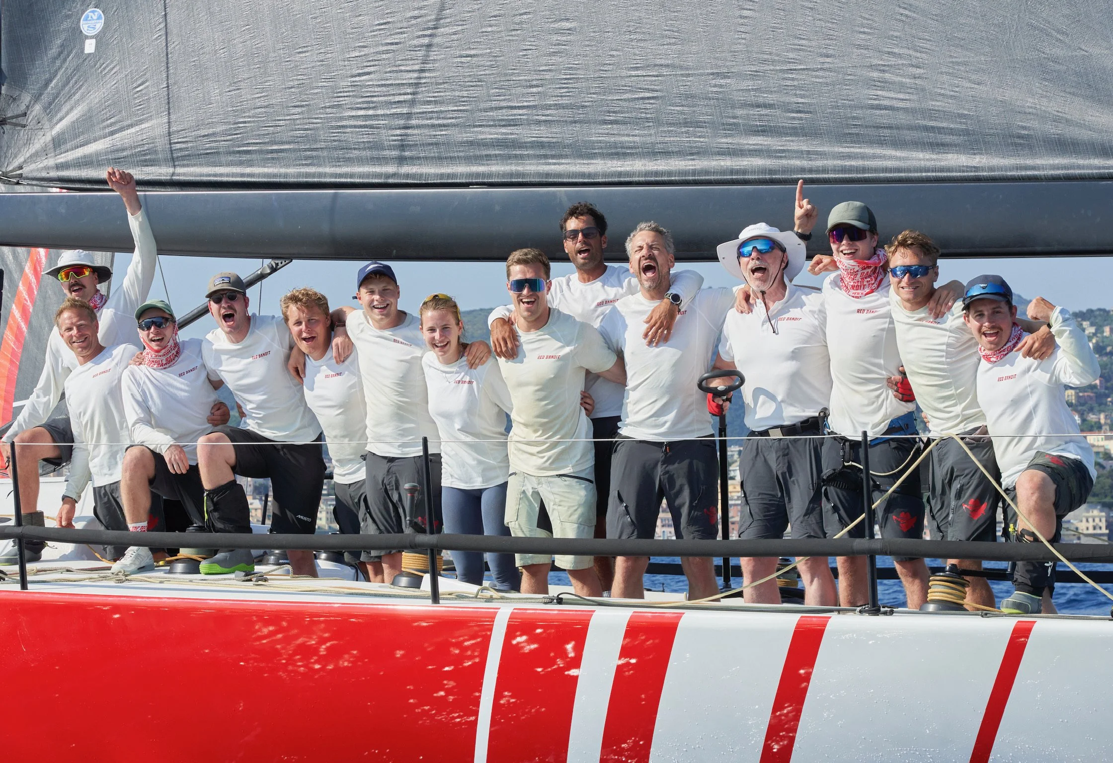 A large group of people on a sailboat celebrating and smiling, with arms around each other, wearing white shirts and sunglasses, with a cityscape in the background.