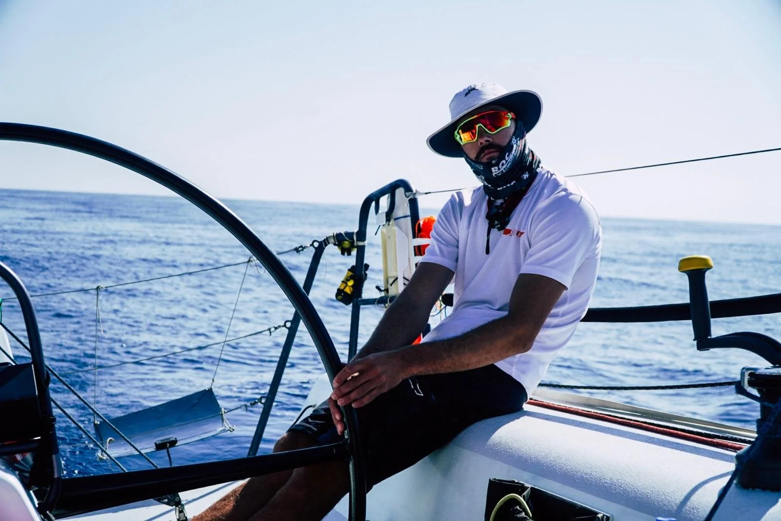 Man in a white shirt and sunglasses on a sailboat, steering the boat while on the open ocean.